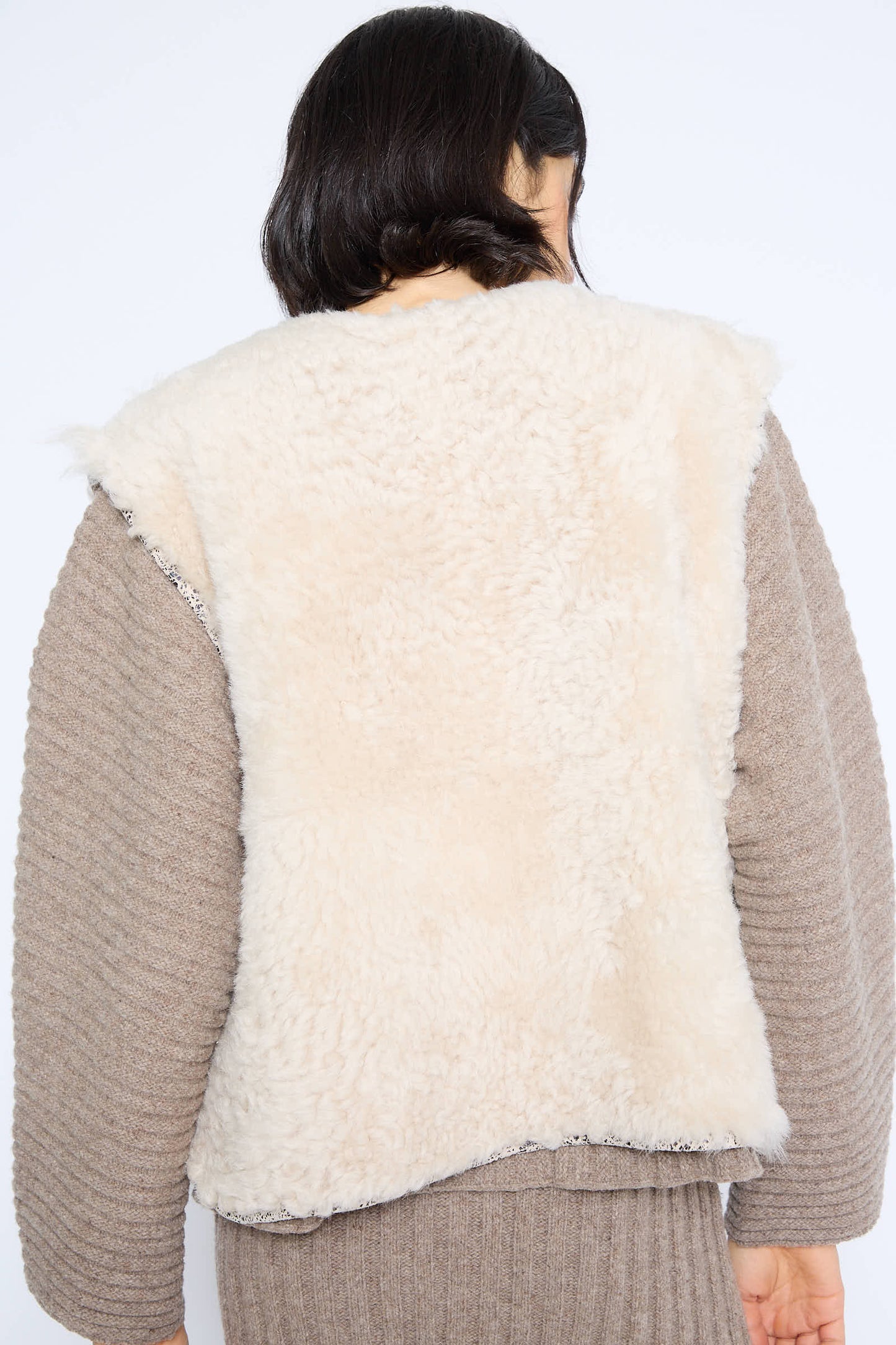 A person with shoulder-length dark hair is facing away, wearing the Milena Silvano Transition Vest in Classic over a taupe sweater, showcasing tonal textures.