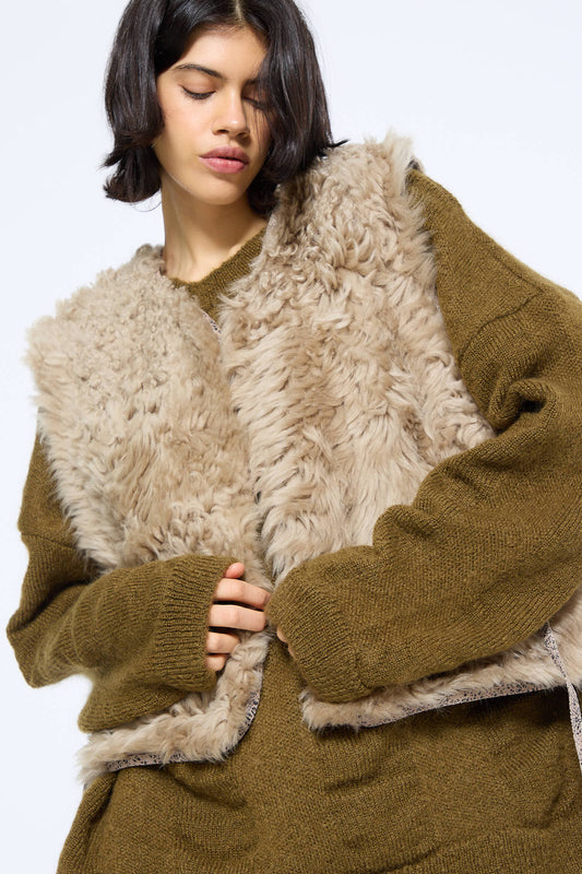 A person wearing the Transition Vest in Reishi by Milena Silvano, a brown furry vest with leather edges, over a green knitted sweater, standing against a plain background.