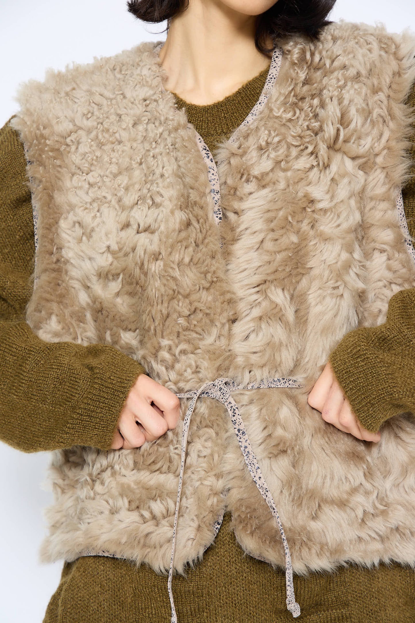 The individual is wearing the Transition Vest in Reishi by Milena Silvano, a beige, fluffy sheepskin vest with leather edges, layered over a green sweater and adjusting the drawstring at the front.