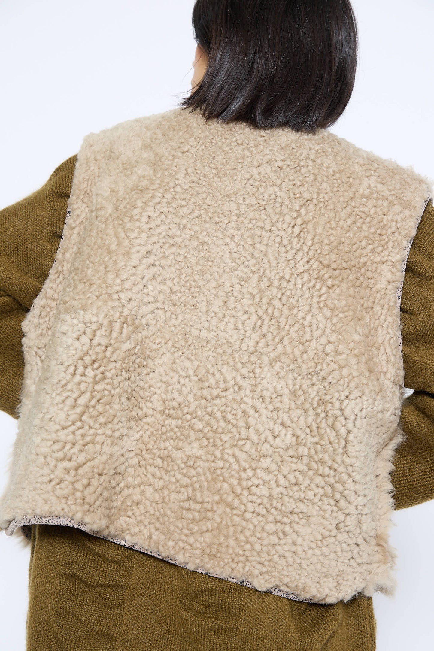 A person wearing the Transition Vest in Reishi by Milena Silvano, a textured, beige sheepskin vest with leather edges, over a green knitted garment, viewed from behind.