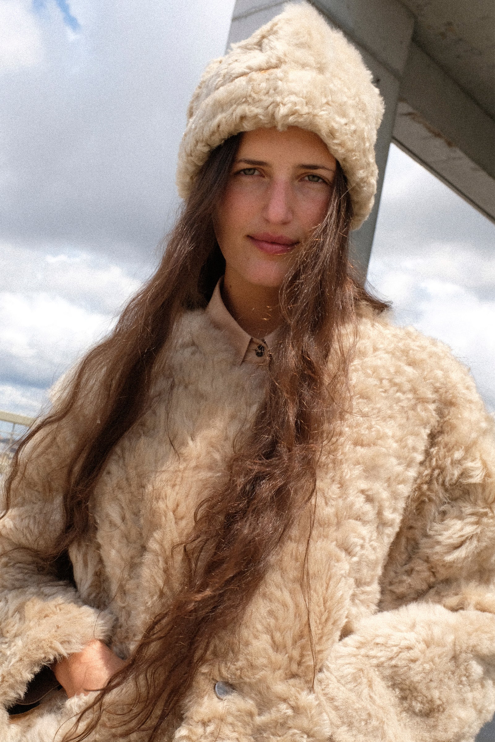 A woman wearing a the Milena Silvano Sheepskin Pixie Hat in Oat, and Sheepskin Reversible Liner in Oat photographed outdoors under a cloudy sky.