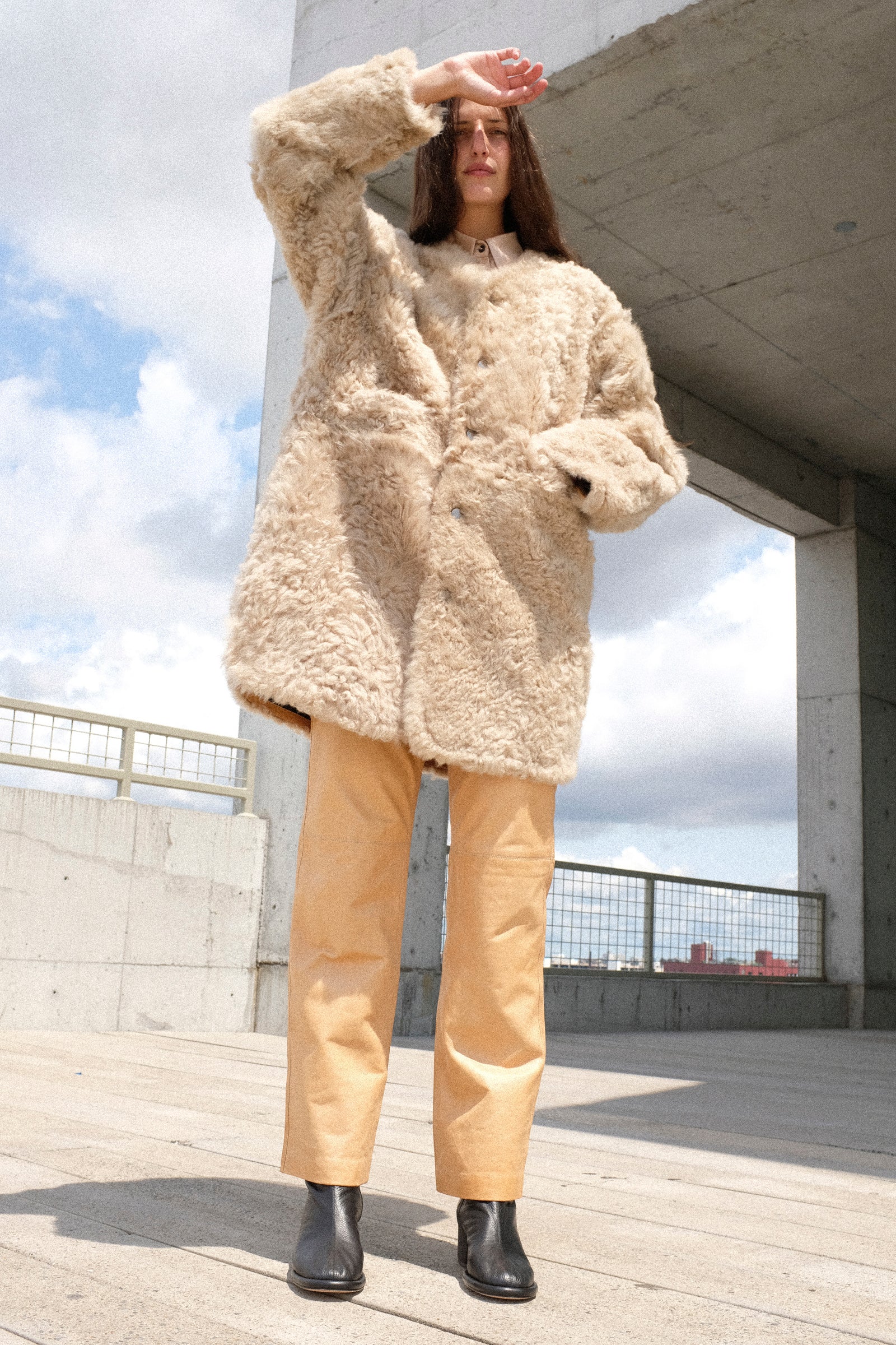 A woman stands outdoors on a concrete structure wearing Milena Silvano's Sheepskin Reversible Liner in Oat and Leather 5 Pocket Trousers in Bare with her hand raised to shield her face from the sun.