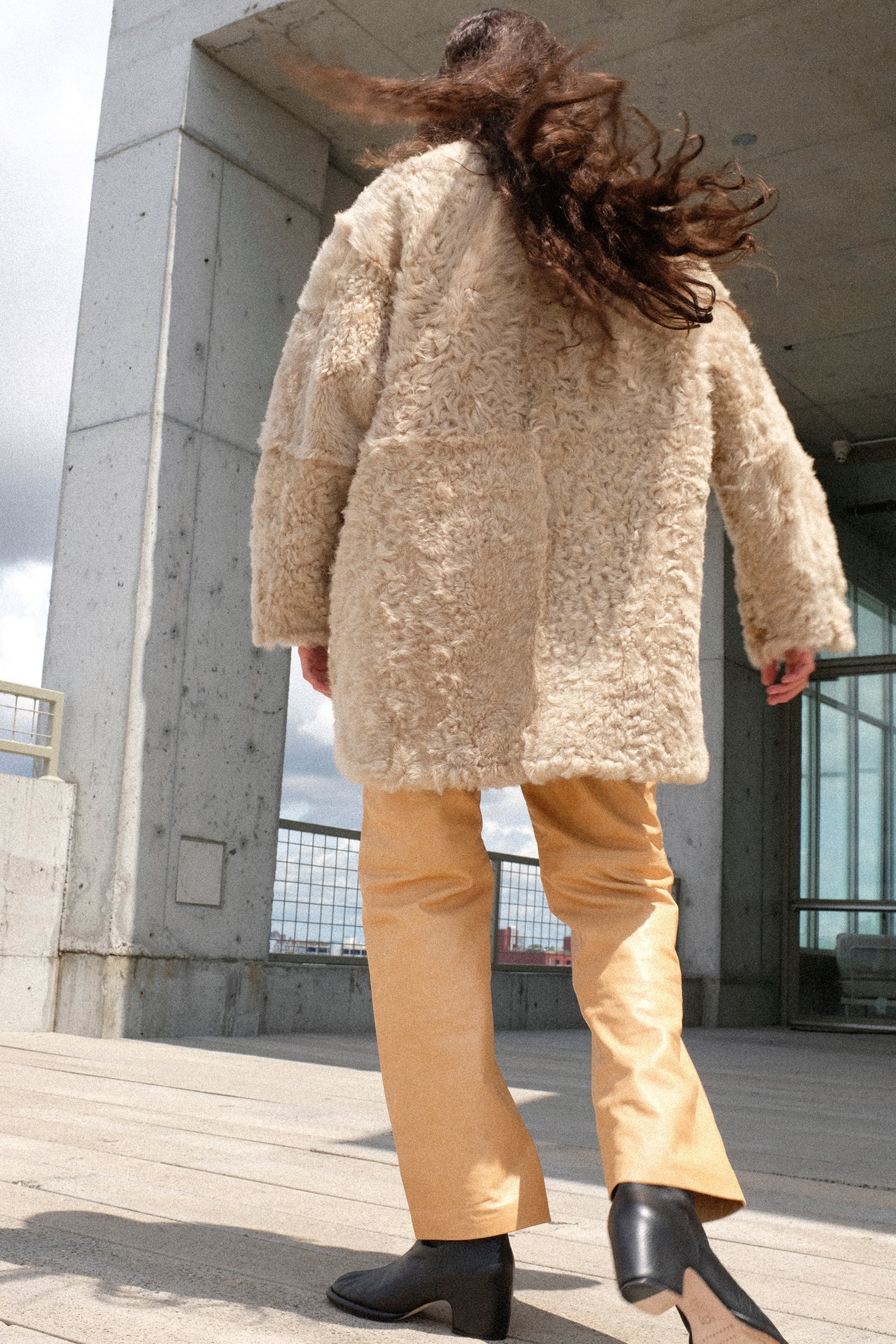 A person shown from behind wearing Milena Silvano Sheepskin Reversible Liner in Oat, and Leather 5 Pocket Trousers in Bare, and black boots while walking on a concrete outdoor walkway.