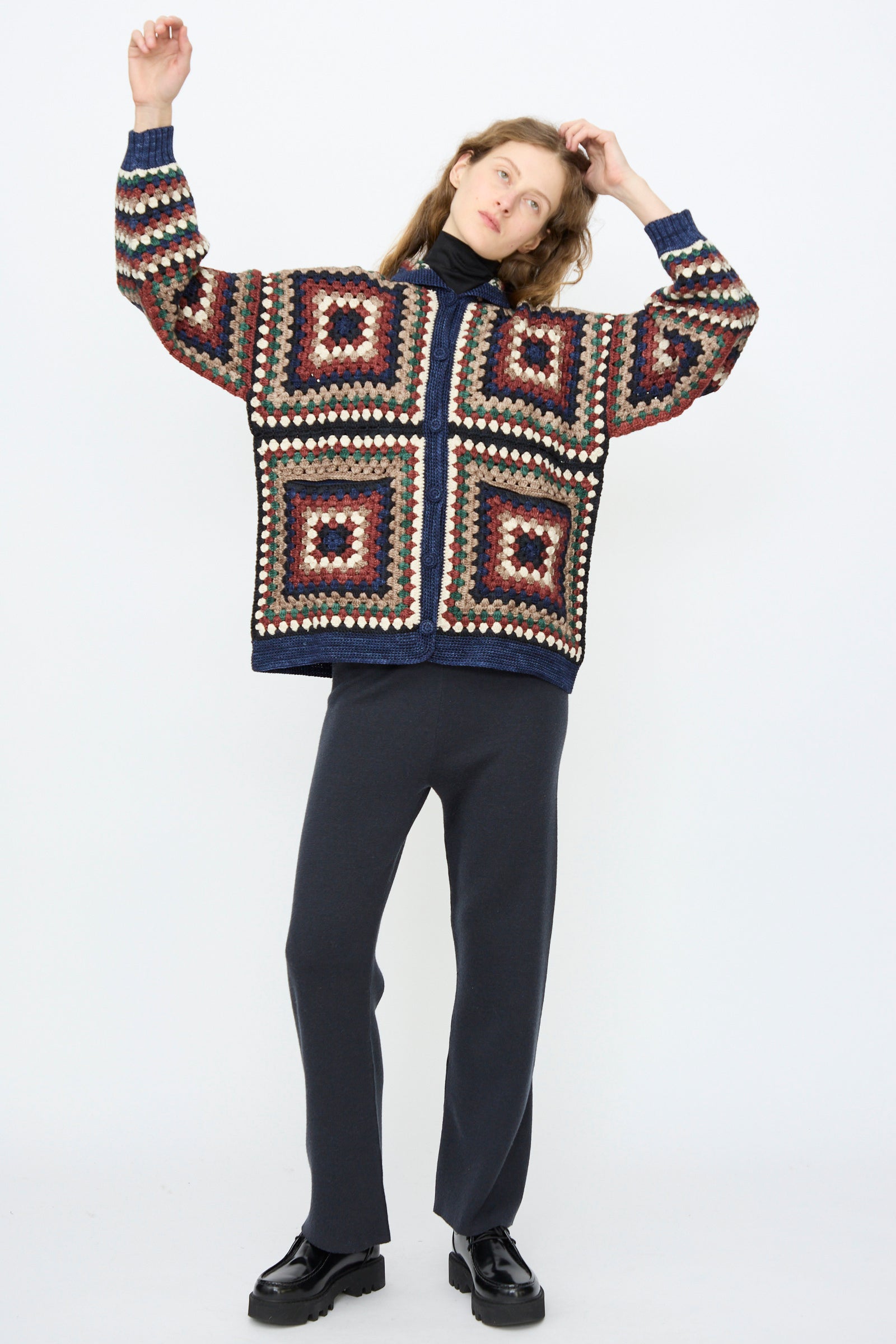 A person stands against a white background wearing the Misha & Puff Crochet Big Square Jacket in Ink over a dark turtleneck and pants with black shoes, arms slightly raised.