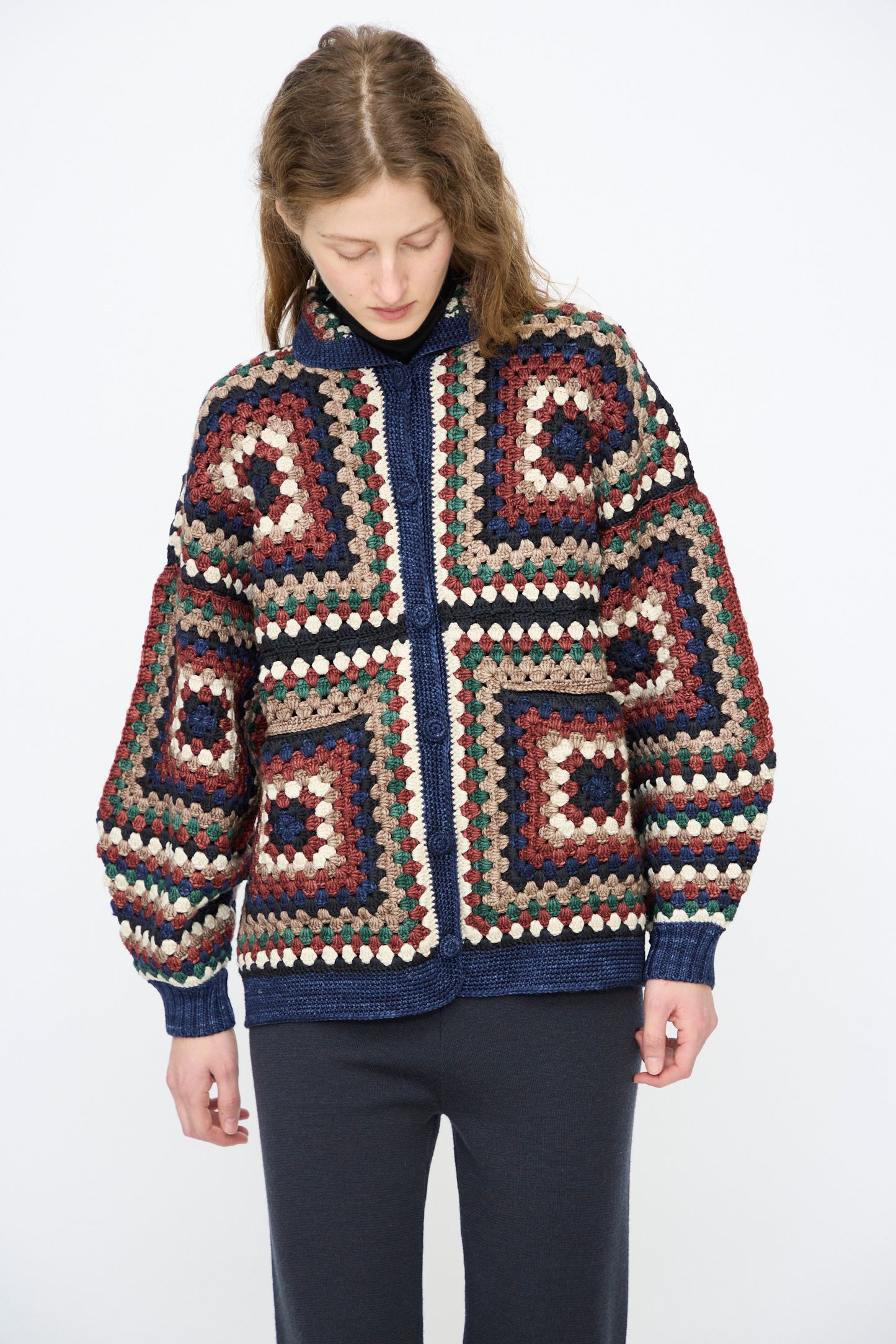 A person wears the Misha & Puff Crochet Big Square Jacket in Ink— a colorful, hand-crocheted cardigan with a zipper—paired with dark pants, looking down against a plain background.