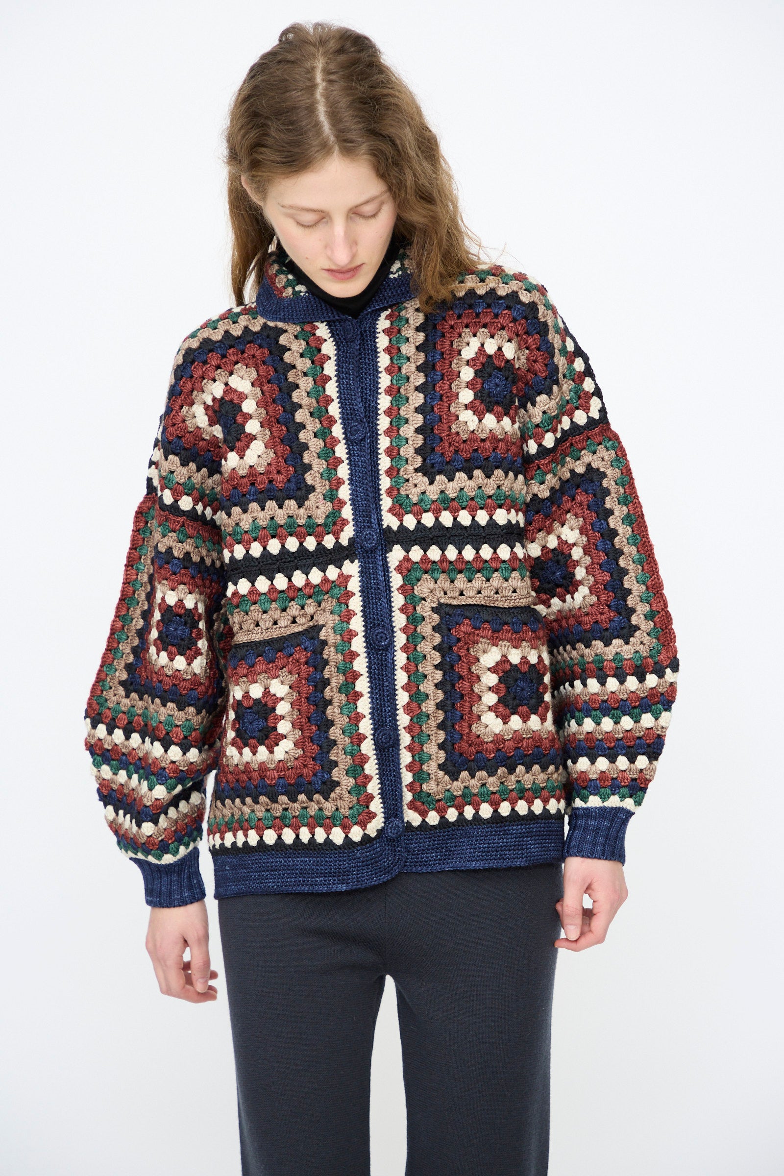 A person wears the Misha & Puff Crochet Big Square Jacket in Ink— a colorful, hand-crocheted cardigan with a zipper—paired with dark pants, looking down against a plain background.