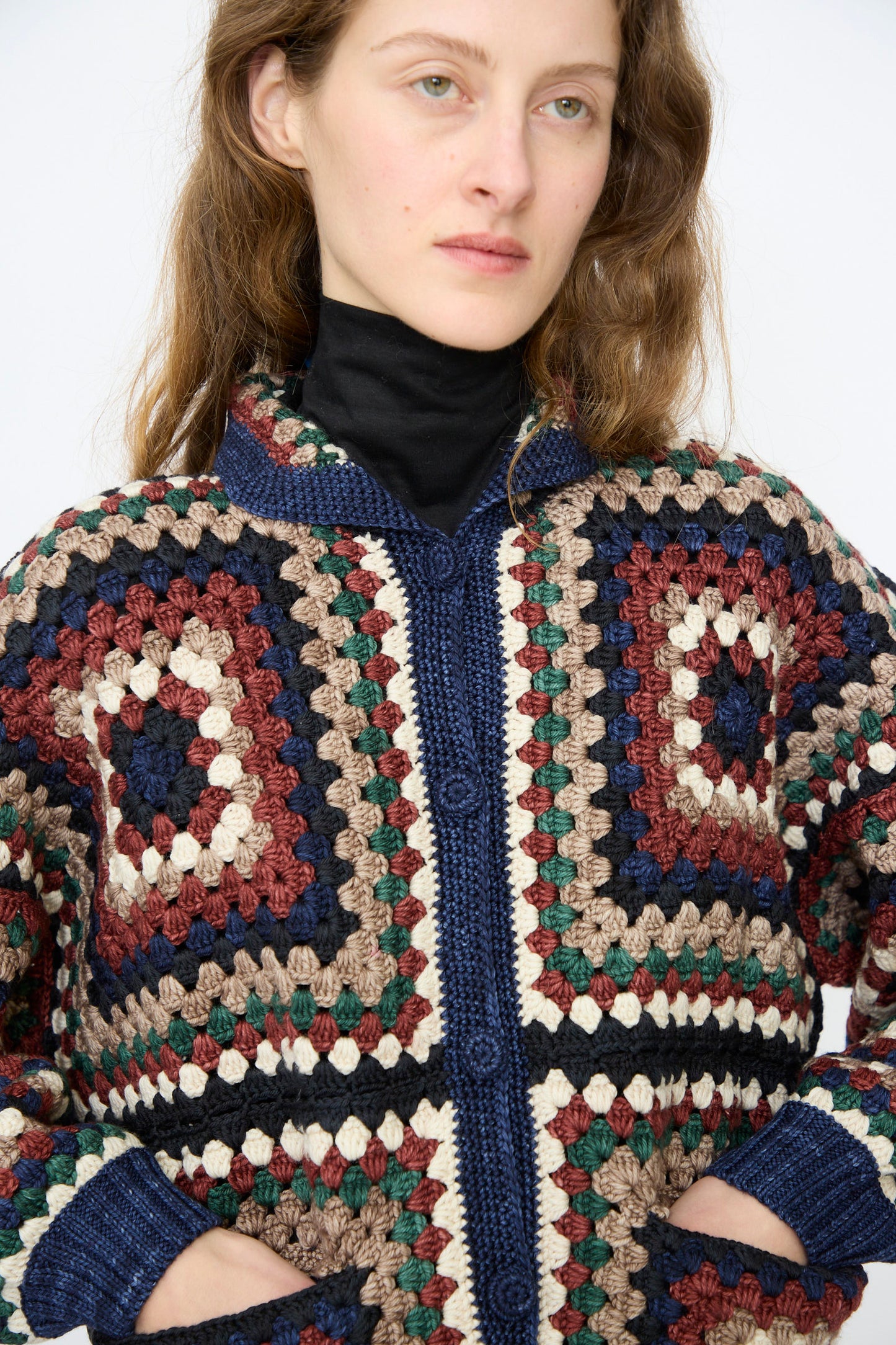 A person models the Misha & Puff Crochet Big Square Jacket in Ink—multicolored, hand-crocheted with classic granny squares—layered over a black turtleneck, hands in pockets, against a plain backdrop.