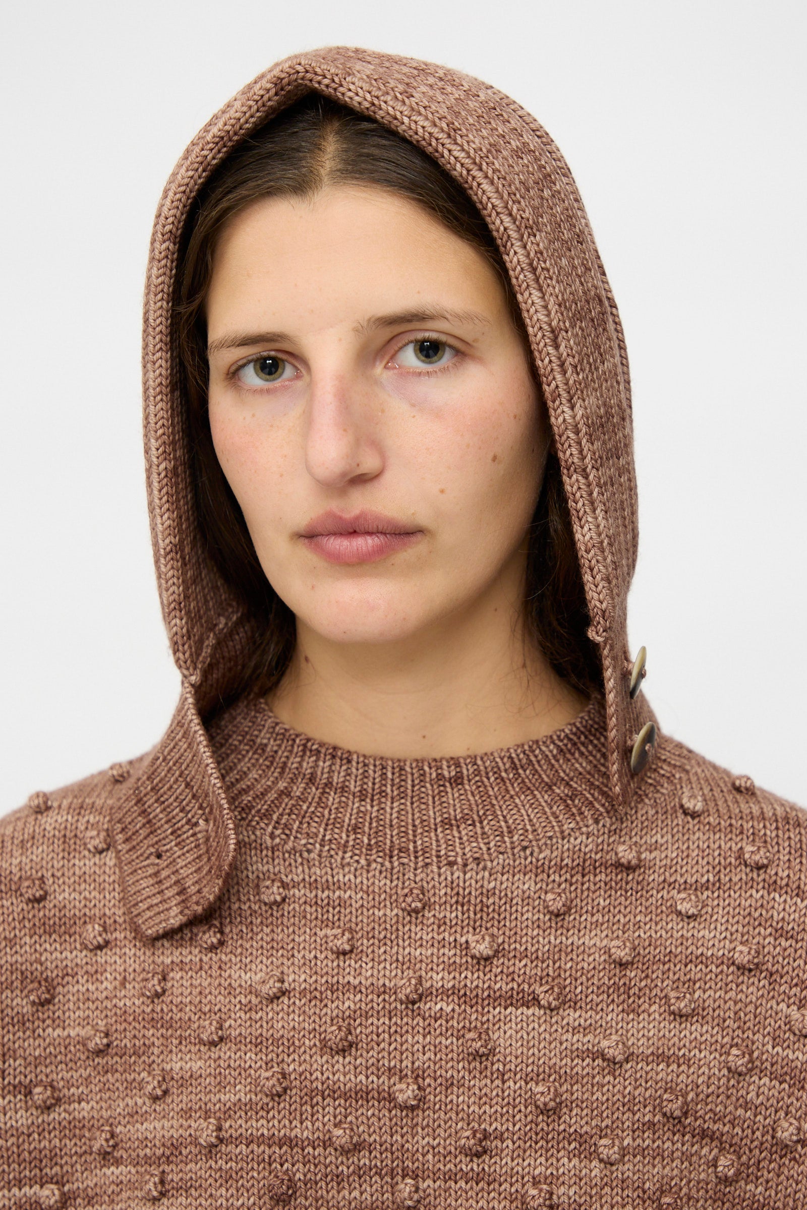 A person wears the Misha & Puff Fisherman Hood in Affogato—a brown knit hooded sweater with horn button closure—while looking directly at the camera against a plain background.