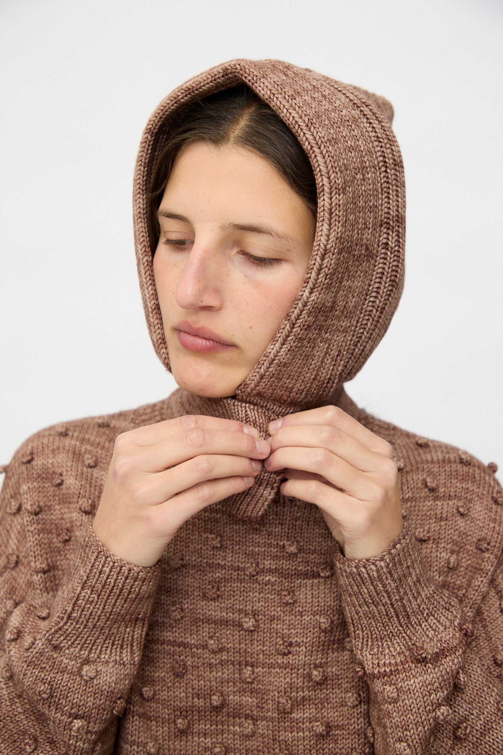 A person wearing the Misha & Puff Fisherman Hood in Affogato, holding the ends of the merino cashmere hood together and looking downwards.