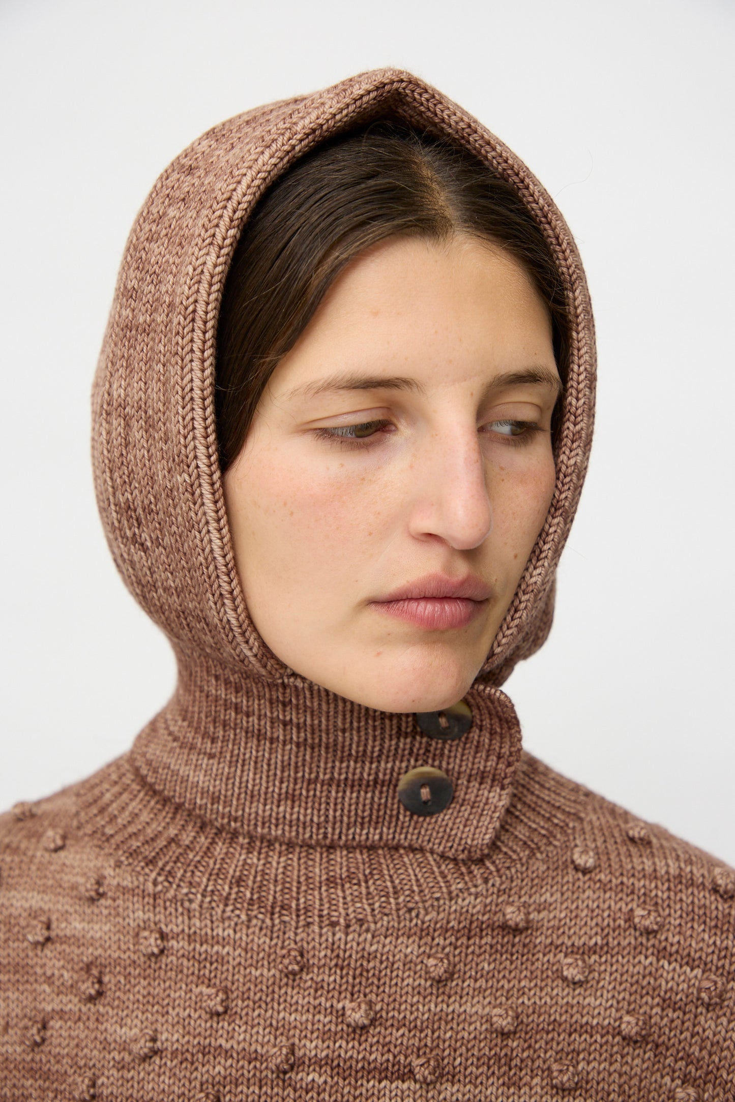 A person wearing the Misha & Puff Fisherman Hood in Affogato—a knitted brown merino cashmere piece with button details—looks to the side against a plain background.