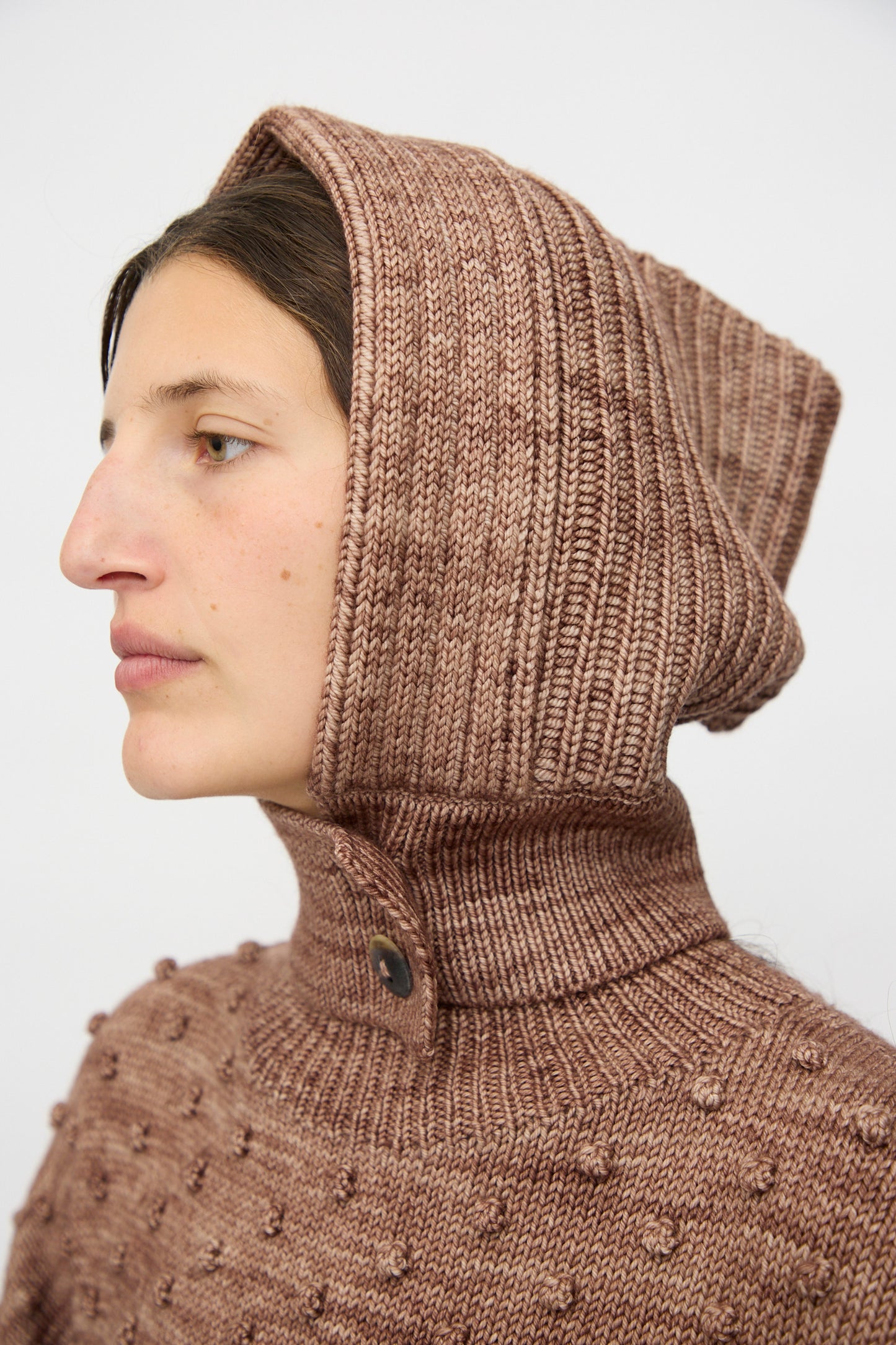 A person wears the Misha & Puff Fisherman Hood in Affogato—a textured brown knit covering head and neck—photographed in profile against a plain background.