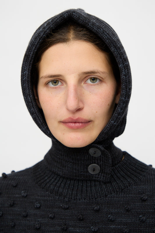 A person with light skin and green eyes wears the Misha & Puff Fisherman Hood in Carbon, featuring two buttons, looking directly at the camera against a plain background.