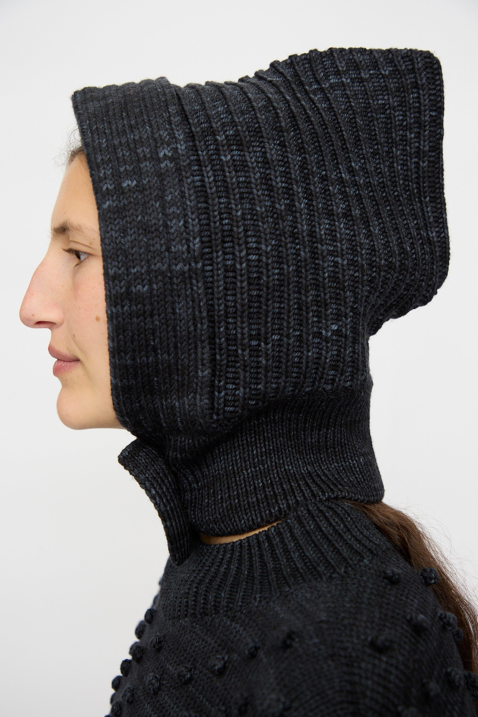 A person in profile wears the Misha & Puff Fisherman Hood in Carbon, a textured black merino cashmere piece covering the head and neck, shown against a plain white background.