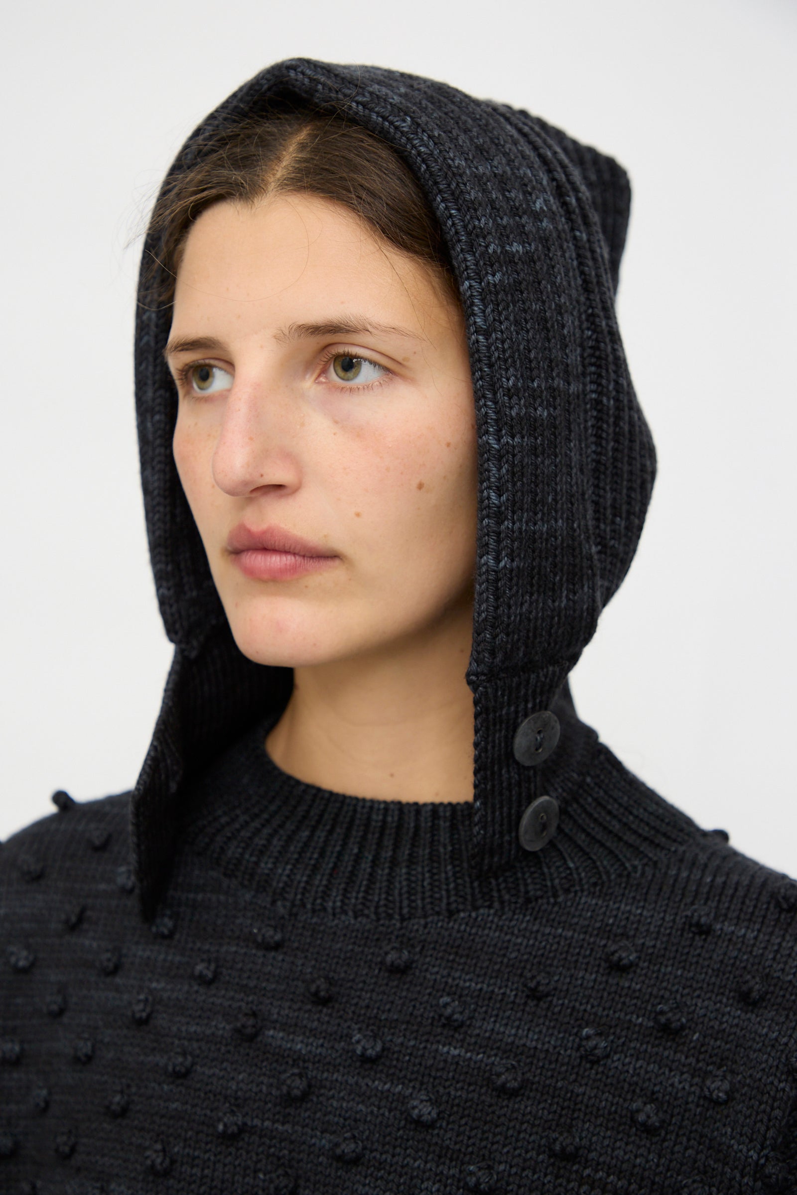 Someone wears the Misha & Puff Fisherman Hood in Carbon, a dark textured knit with an attached merino cashmere hood and two buttons, looking off to the side against a plain background.