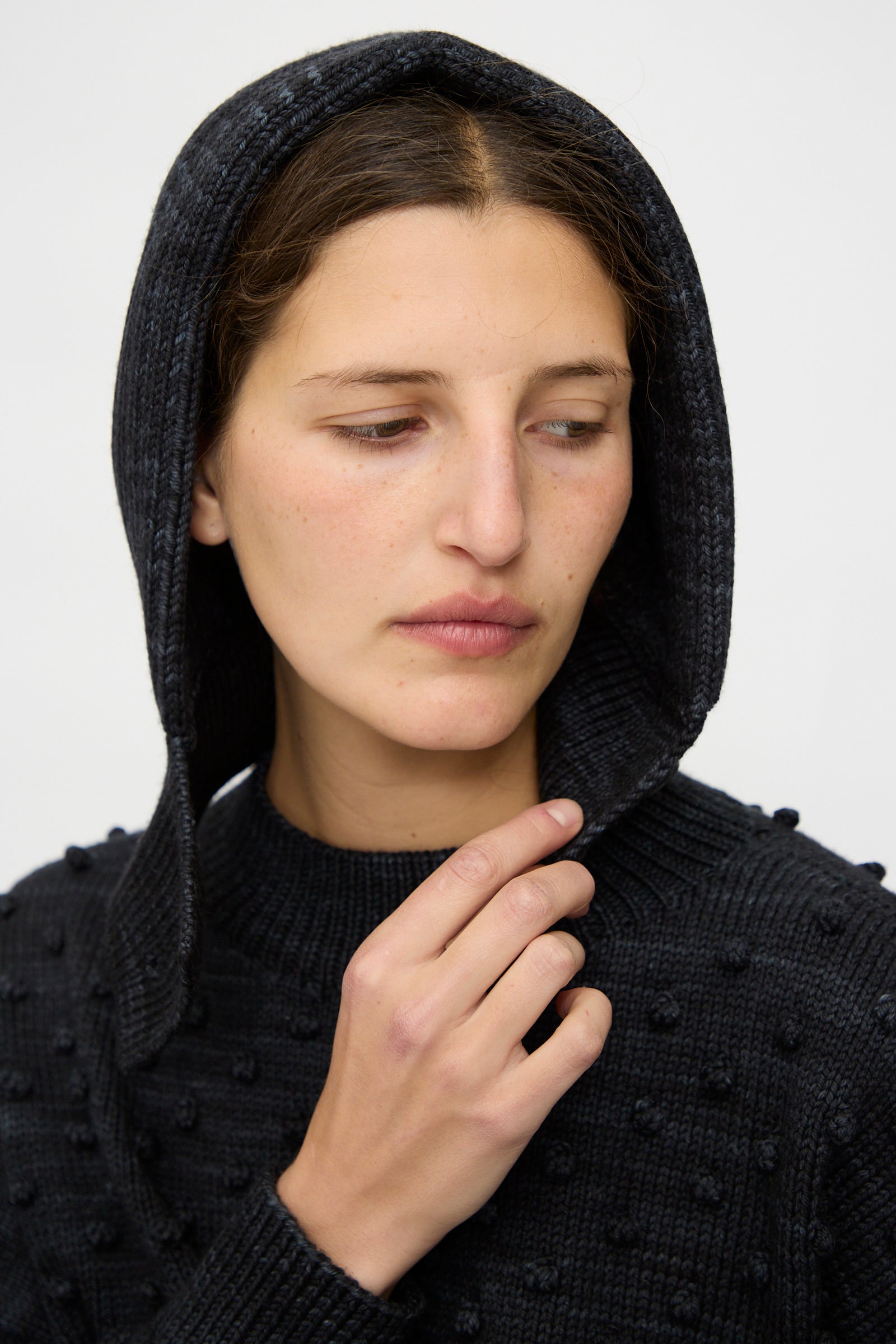 A person wearing the Fisherman Hood in Carbon by Misha & Puff looks down and to the side while holding the edge of the hood.