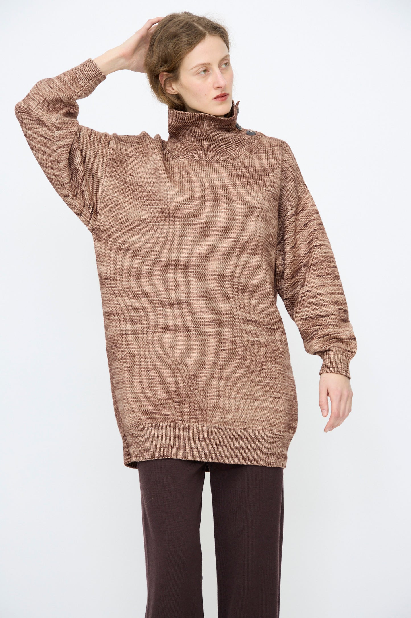 A person wearing the Hutton Dress in Affogato by Misha & Puff stands against a plain background, one hand raised to their head.