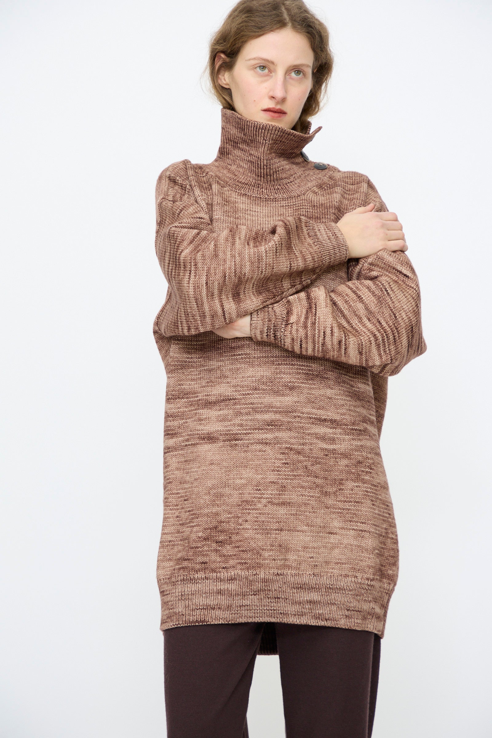 A person wears the Hutton Dress in Affogato by Misha & Puff, a loose brown hand-dyed turtleneck mini dress, paired with dark pants, standing with arms crossed against a plain white background.