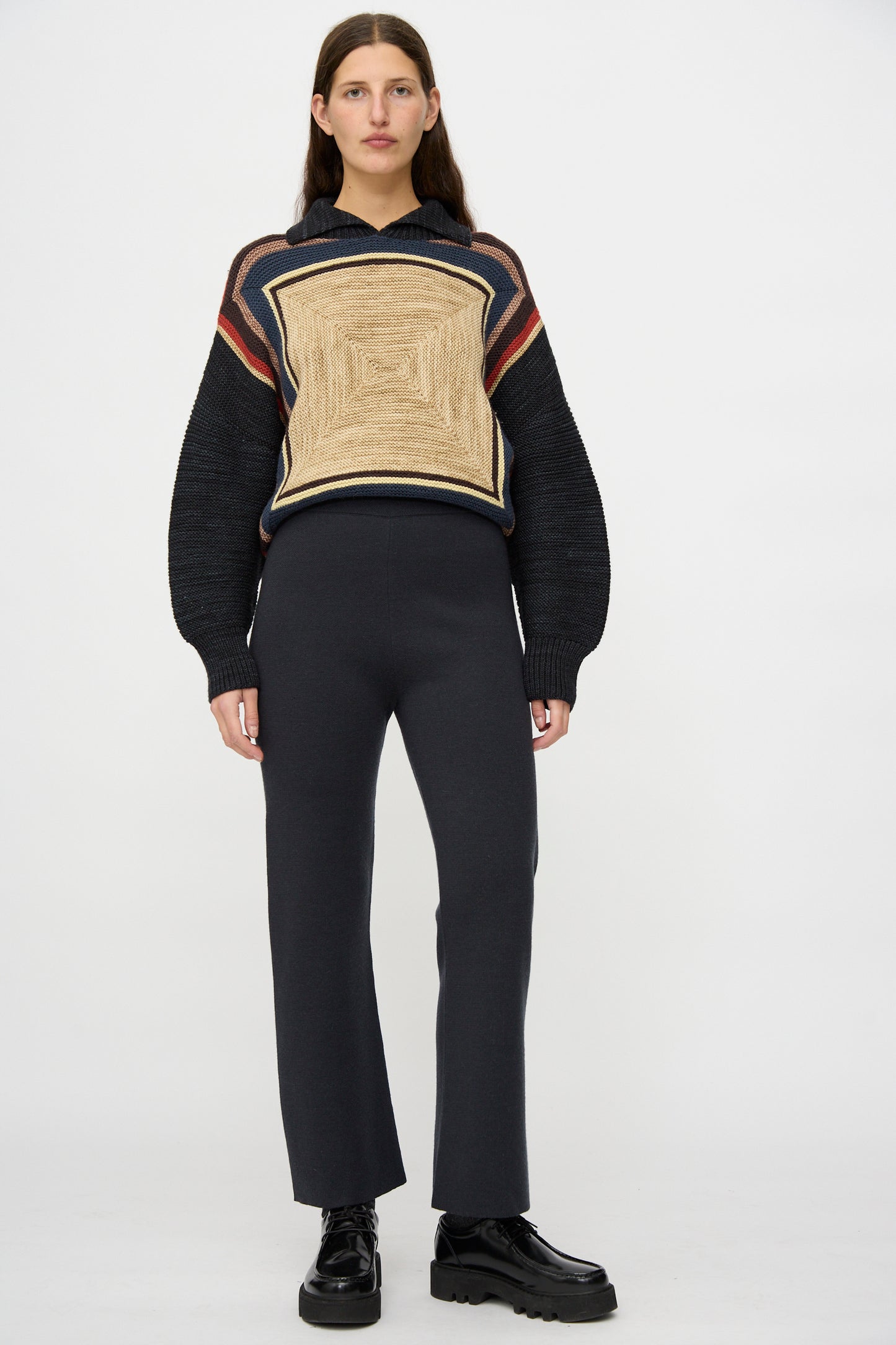 A person stands facing forward against a plain white background, wearing the Misha & Puff Izzy Pant in Carbon, styled with a geometric-patterned sweater and black platform shoes.
