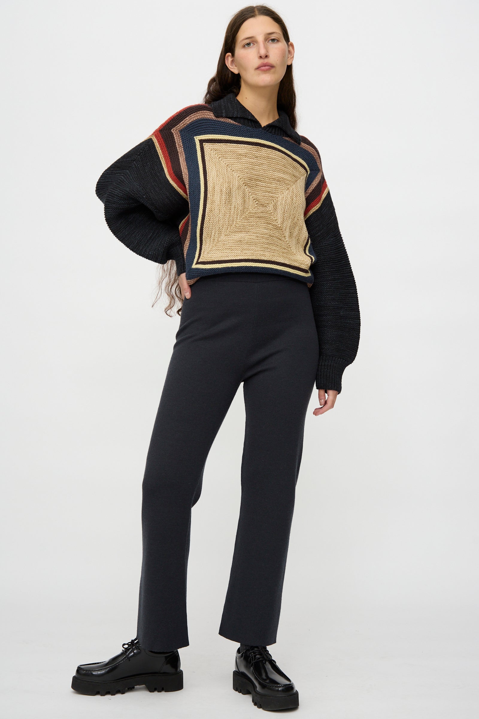 A woman stands against a plain background wearing the Misha & Puff Izzy Pant in Carbon, styled with a patterned sweater and black platform shoes.