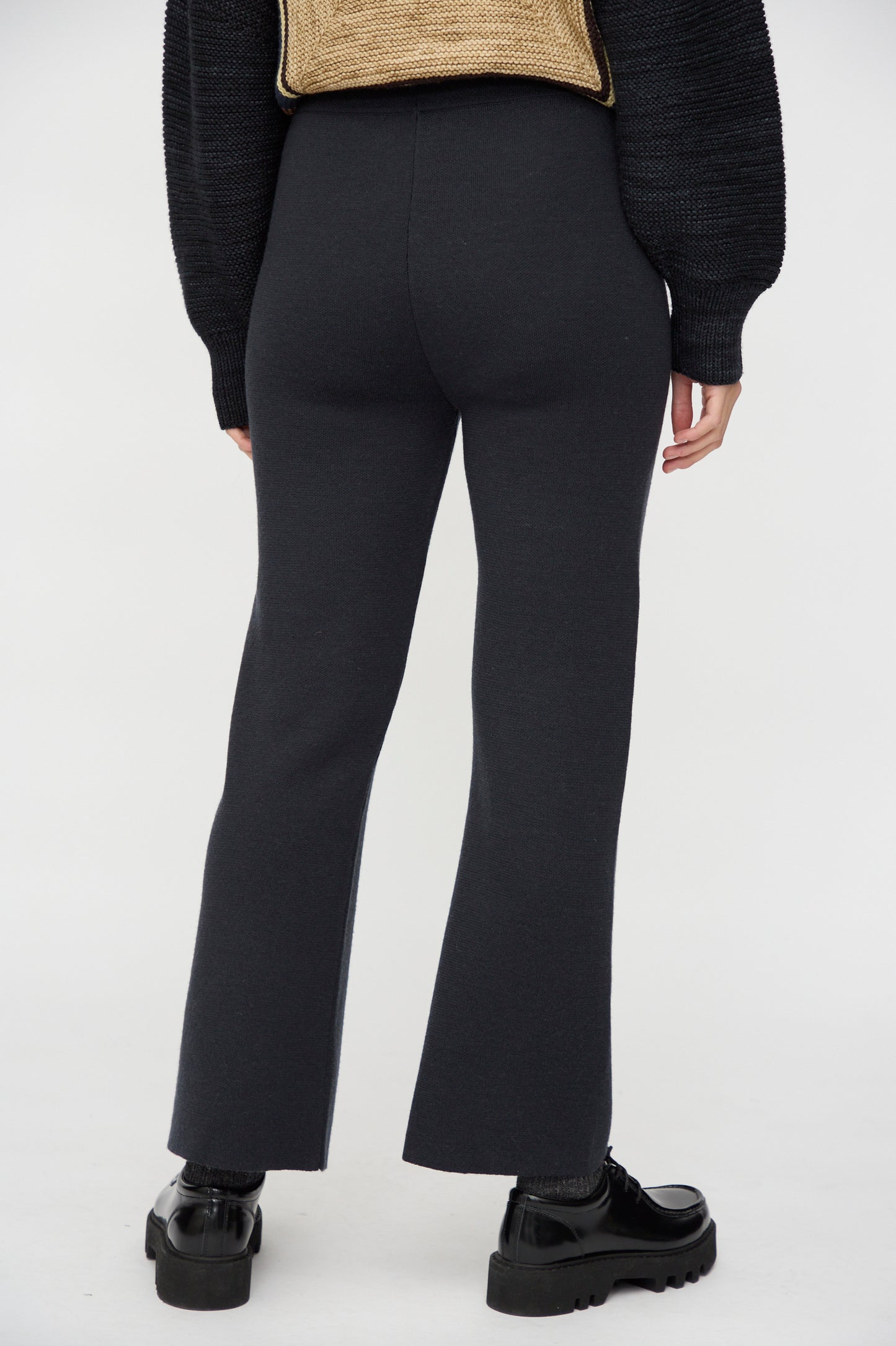 A person stands with their back to the camera against a white background, wearing Misha & Puff's Izzy Pant in Carbon and a black textured sweater paired with black shoes.