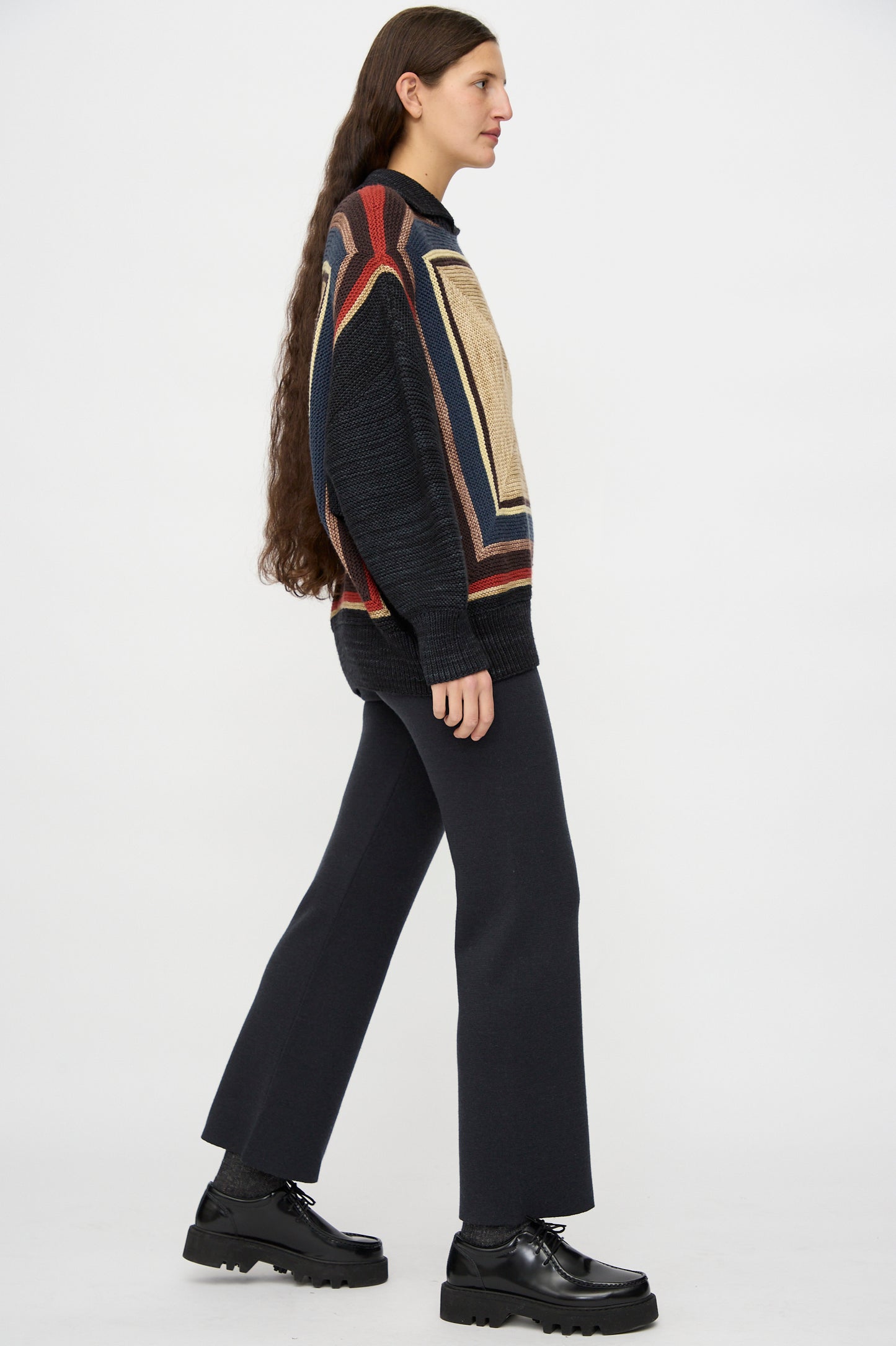 A person with long brown hair, wearing a patterned sweater, Misha & Puff's Izzy Pant in Carbon, and black platform shoes stands in profile against a plain white background.