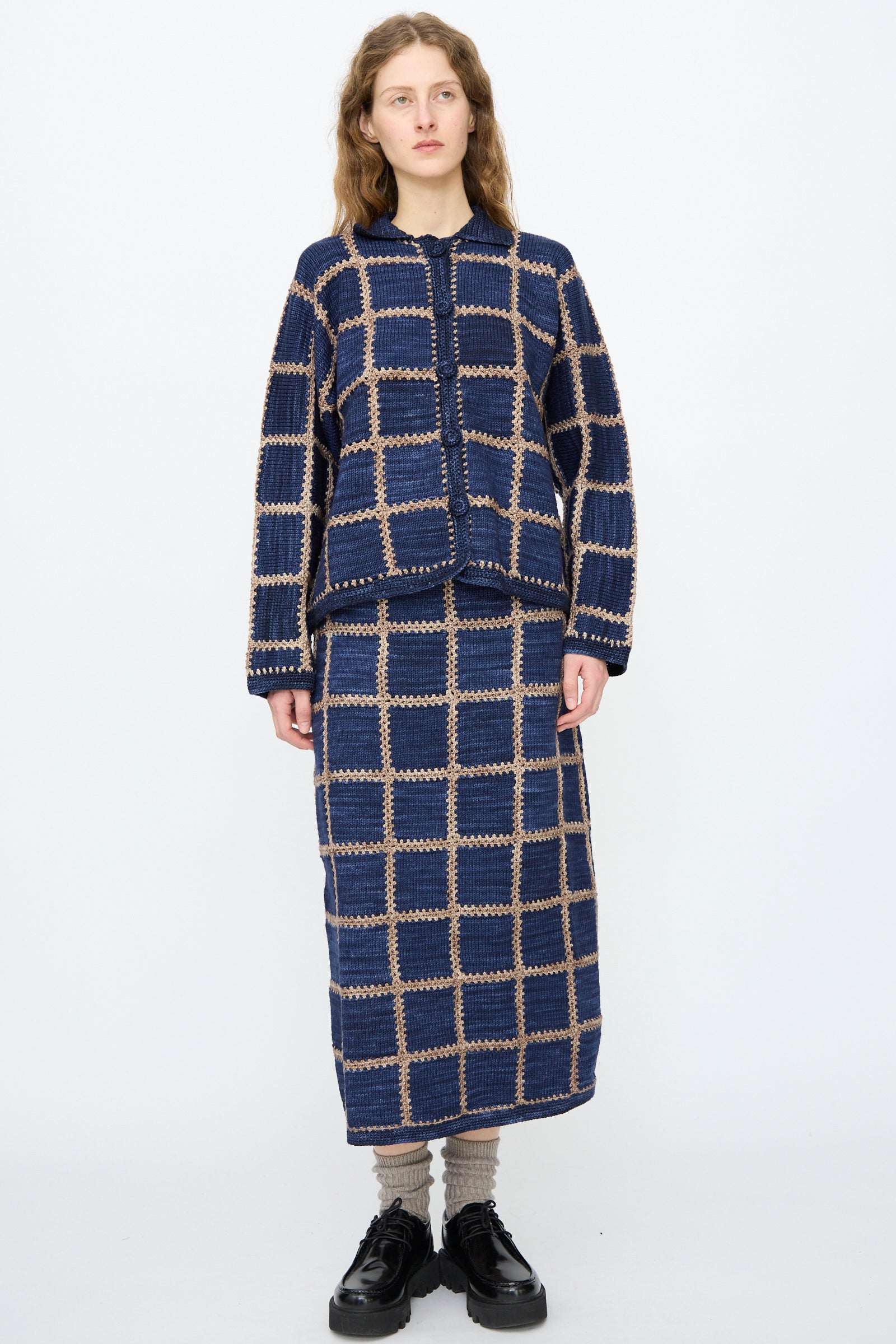 A person faces forward in a Misha & Puff Ladder Square Cardigan in Ink with a matching long skirt, grey socks, and black shoes, standing against a plain white background.