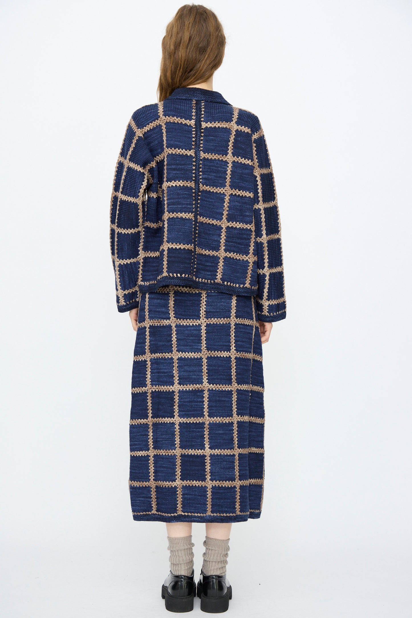 A person with long hair stands facing away, wearing Misha & Puff's Ladder Square Cardigan in Ink over a blue and beige plaid jacket, a matching skirt, black shoes, and beige socks.