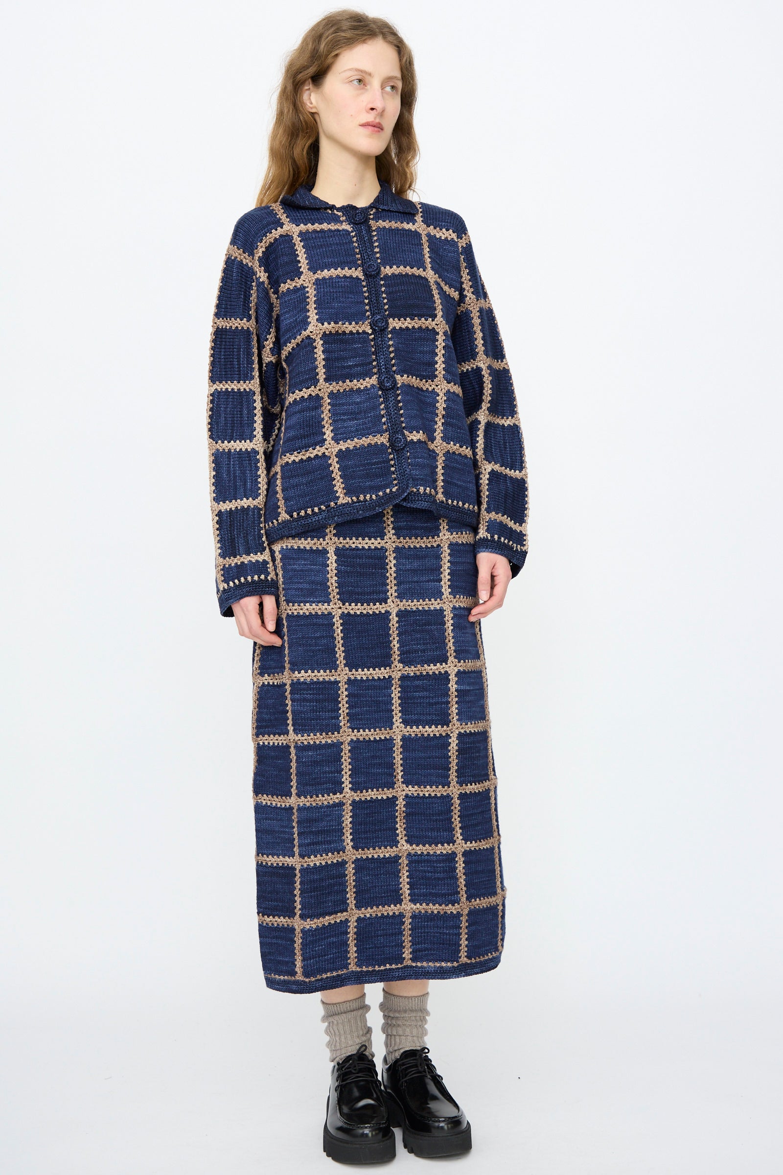 A woman wears the Misha & Puff Ladder Square Cardigan in Ink, styled with a matching checkered skirt, beige socks, and black platform shoes, standing against a plain white background.