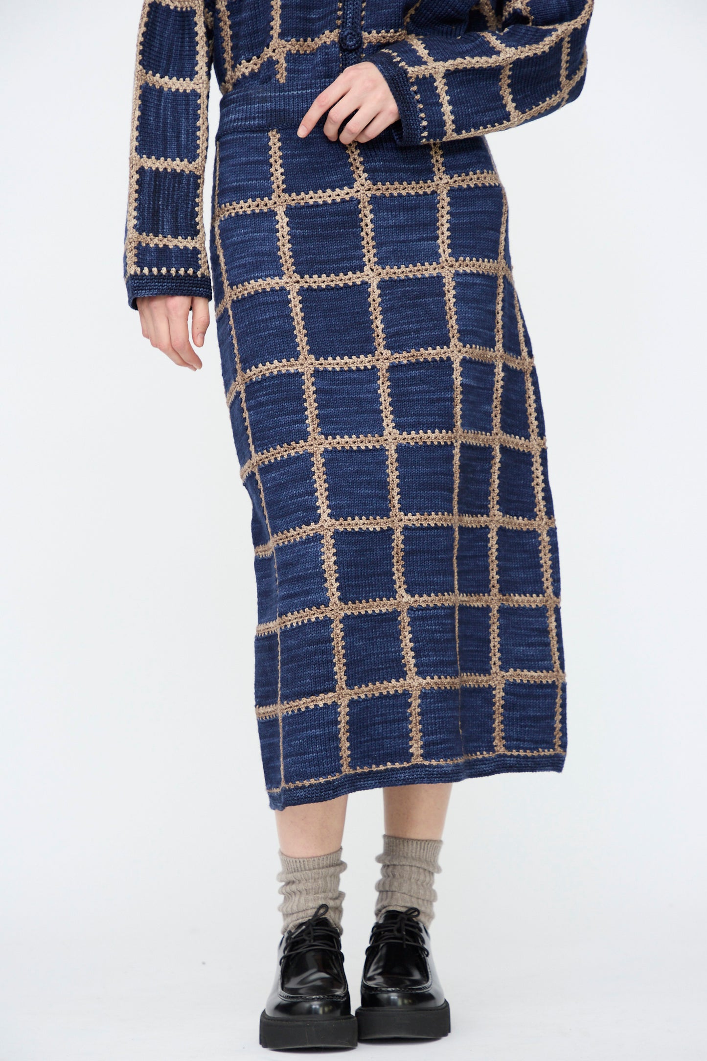 A person wears the Misha & Puff Ladder Square Skirt in Ink with a matching top with hand-crocheted details, gray socks, and black platform shoes, standing against a plain background.