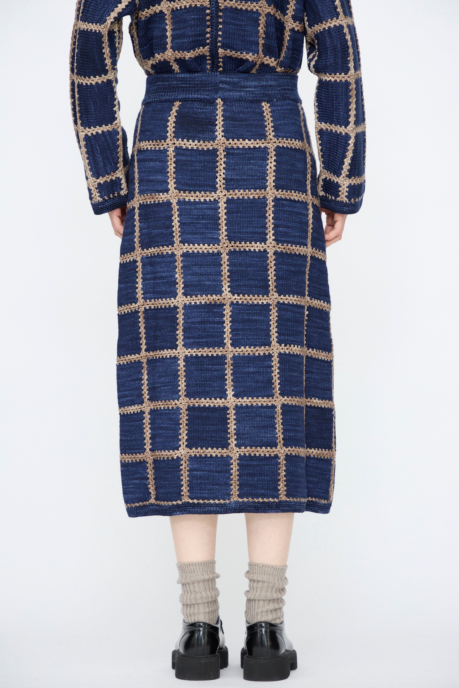 A person stands facing away, wearing the Misha & Puff Ladder Square Skirt in Ink with a matching top, subtle knit patchwork, gray socks, and black shoes against a plain white background.