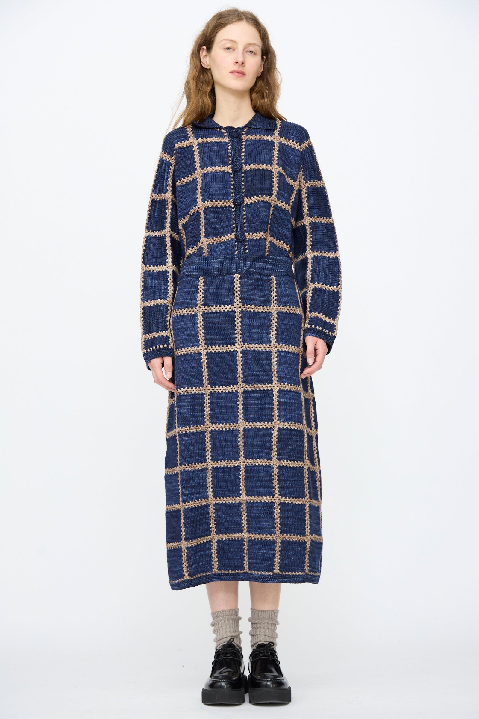 A woman stands against a plain background wearing the Misha & Puff Ladder Square Skirt in Ink, paired with beige socks and black platform shoes.