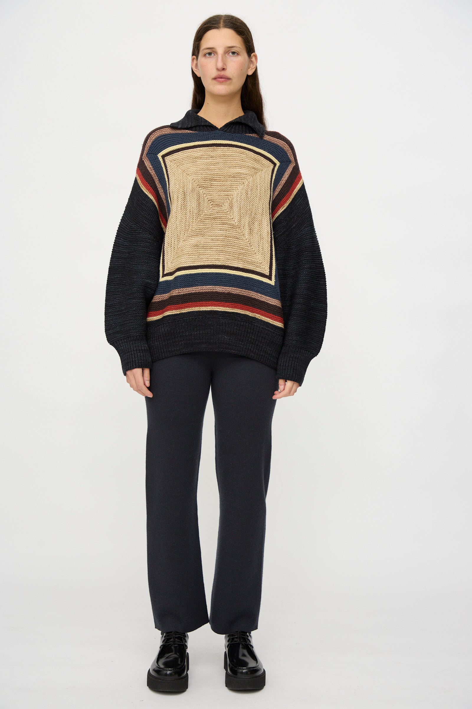 A person stands against a white background wearing the Misha & Puff Log Cabin Sweater in Carbon, paired with dark pants and black shoes.