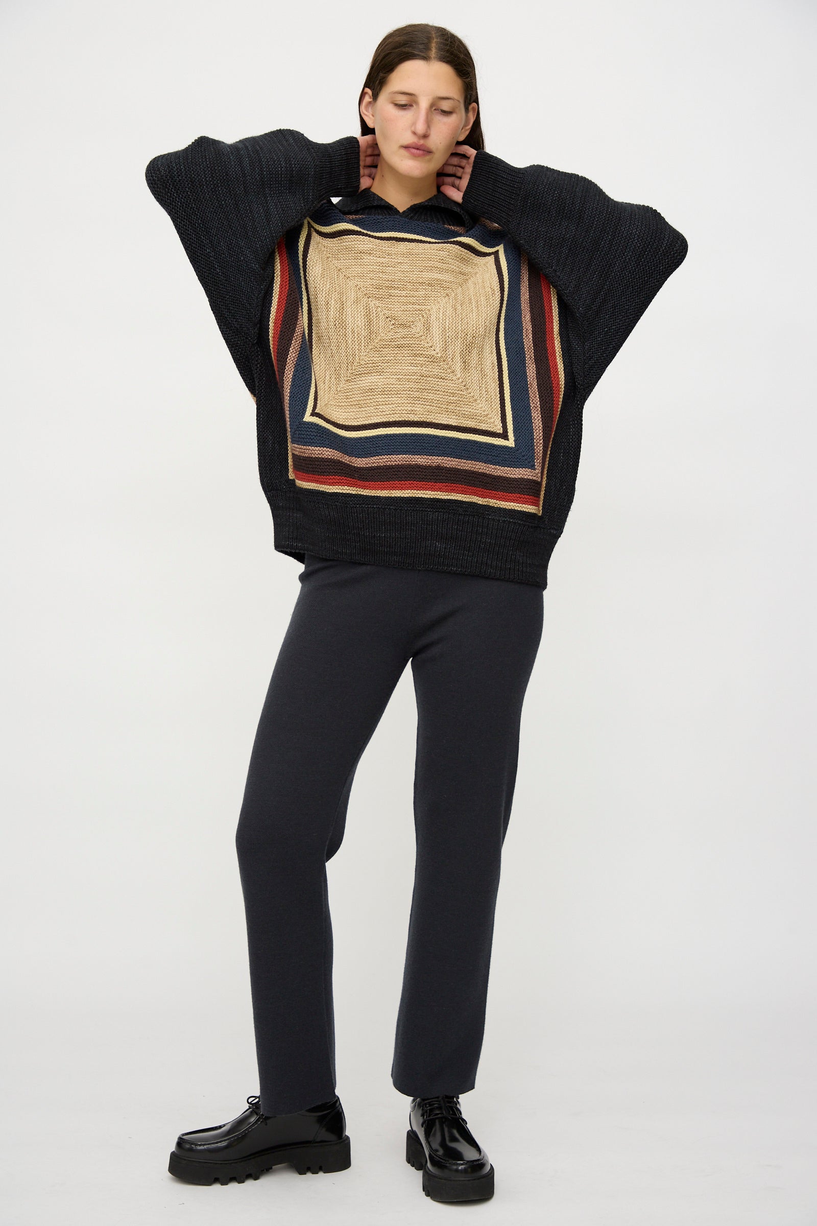 A person with eyes closed wears the Misha & Puff Log Cabin Sweater in Carbon—featuring a geometric square pattern and a loose fit—paired with dark pants and black platform shoes, standing against a plain background.