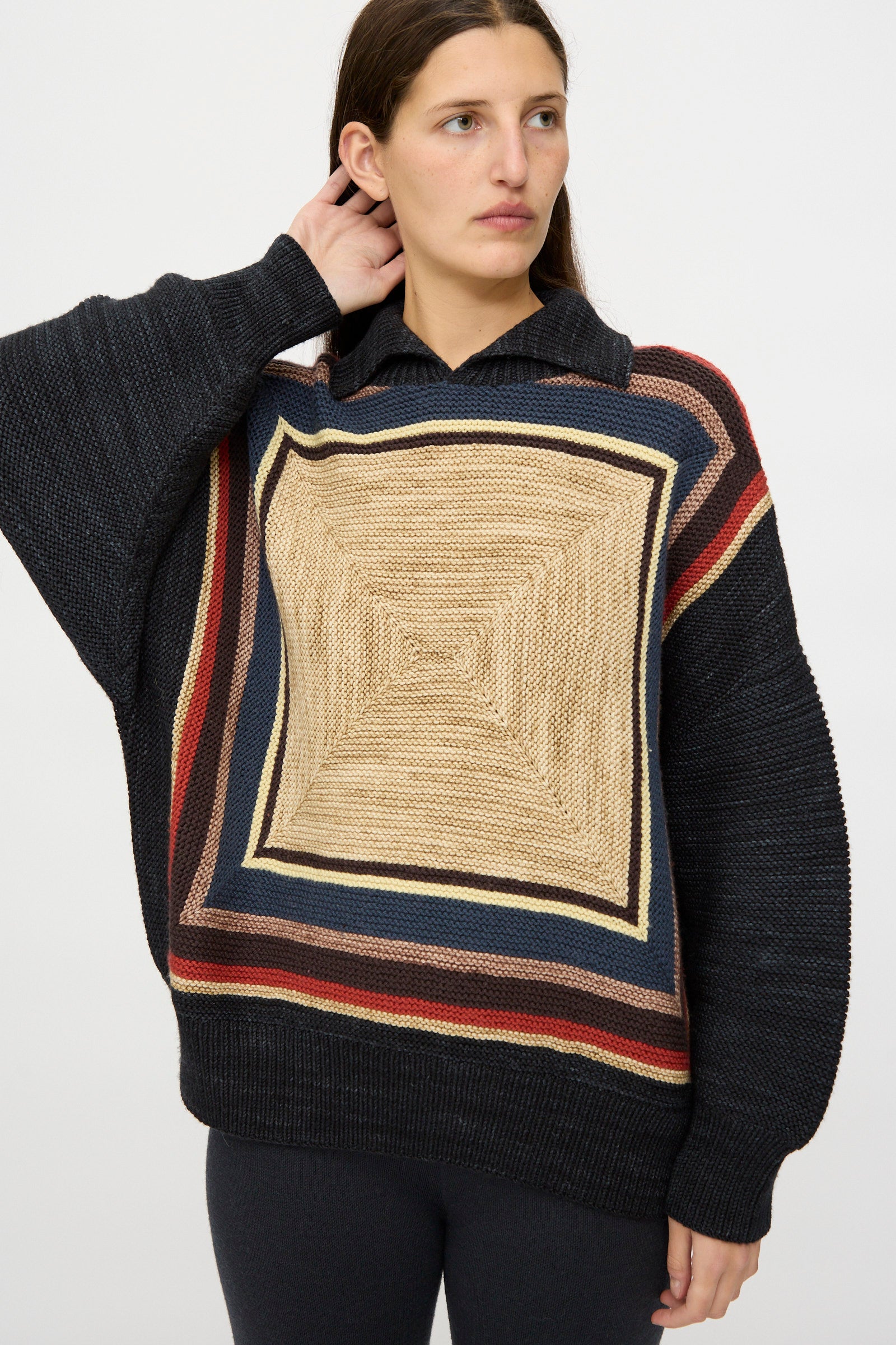 A woman wears the Misha & Puff Log Cabin Sweater in Carbon—featuring a loose fit, large square geometric pattern in beige, red, and navy, crafted from soft merino wool in Peru—posed against a plain background.