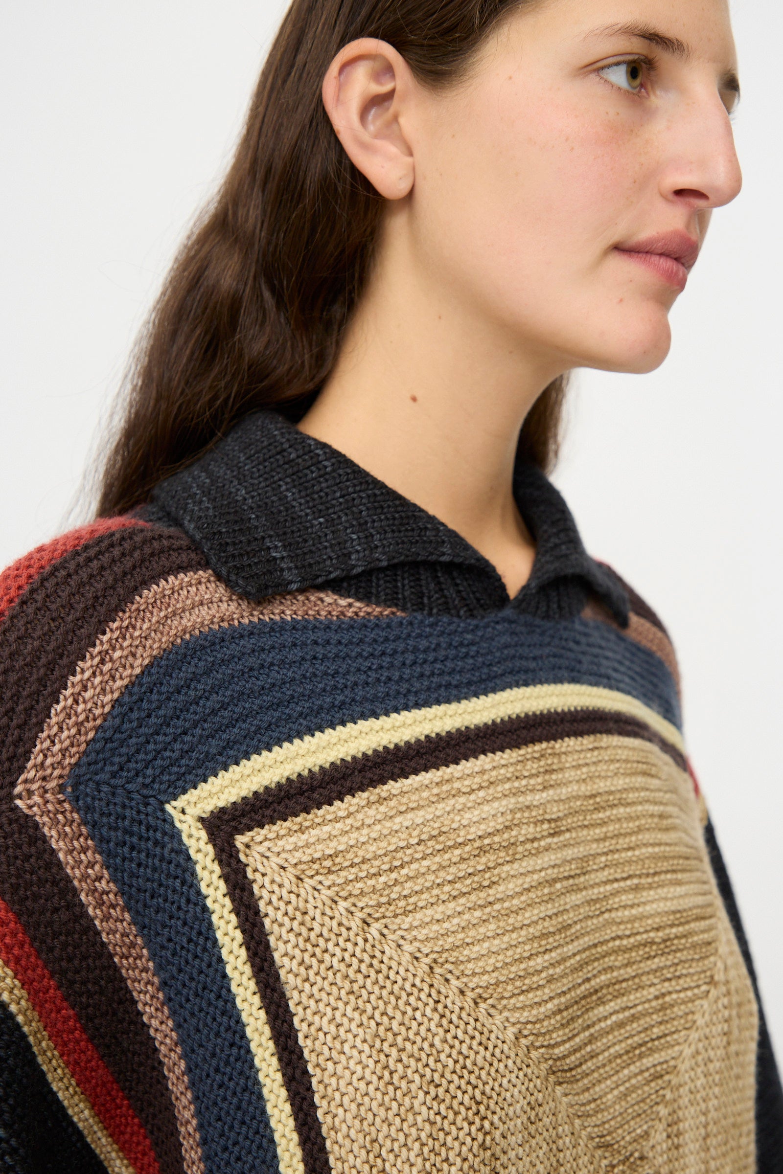 A woman with long dark hair wears the Misha & Puff Log Cabin Sweater in Carbon—a multicolored, geometric knit—over a black collared shirt, looking to the side against a plain background.
