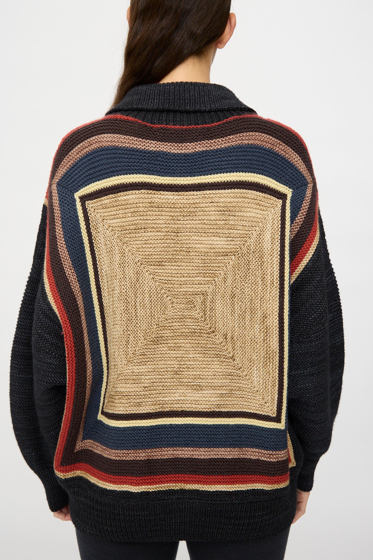 Someone wearing the Misha & Puff Log Cabin Sweater in Carbon—a Peruvian-made sweater with a black base, large beige geometric square, and multicolored stripes on the back—faces away from the camera.