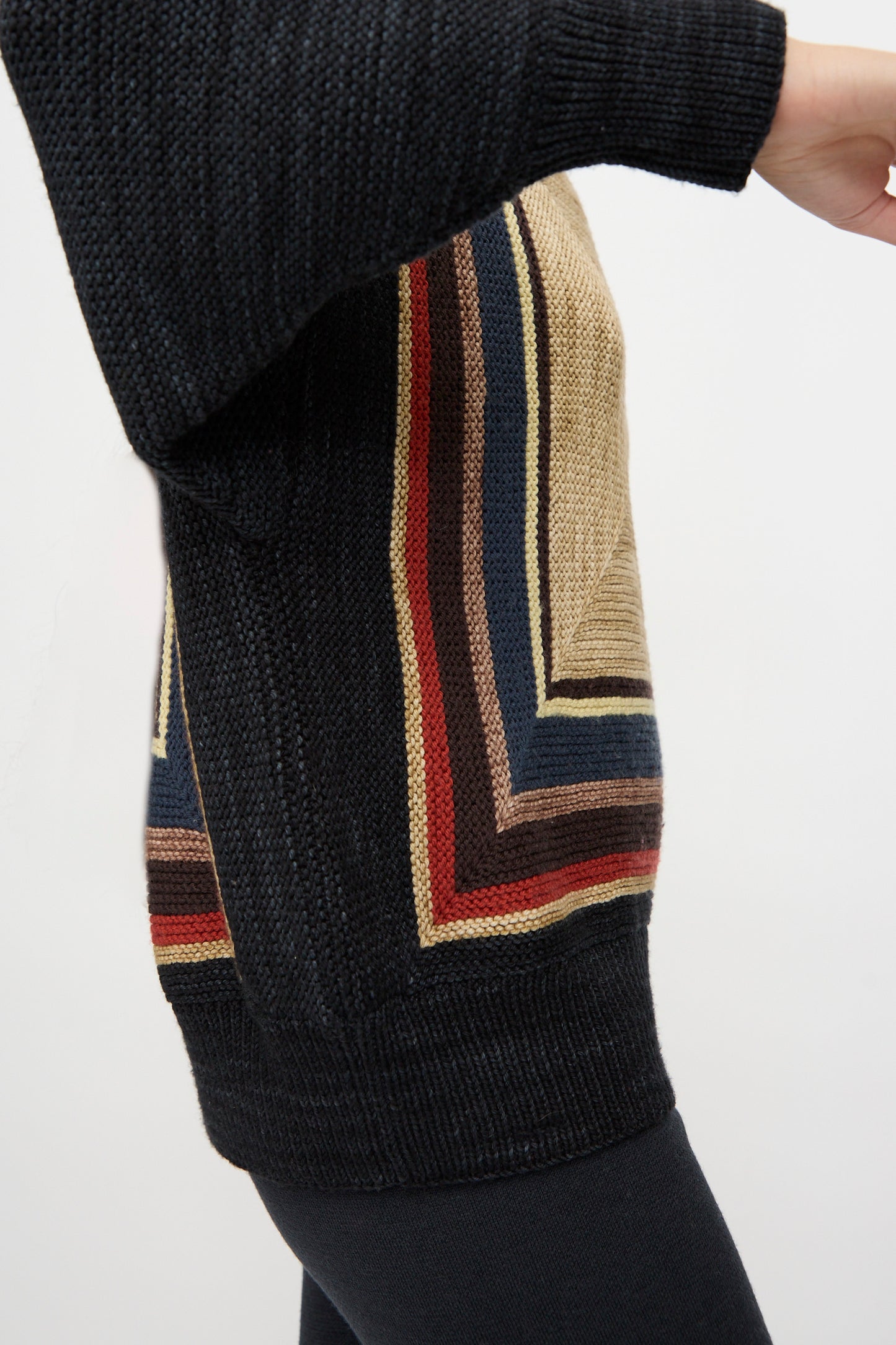 A person wearing the Misha & Puff Log Cabin Sweater in Carbon, featuring black sleeves and multicolored geometric stripes, made in Peru, paired with black pants.