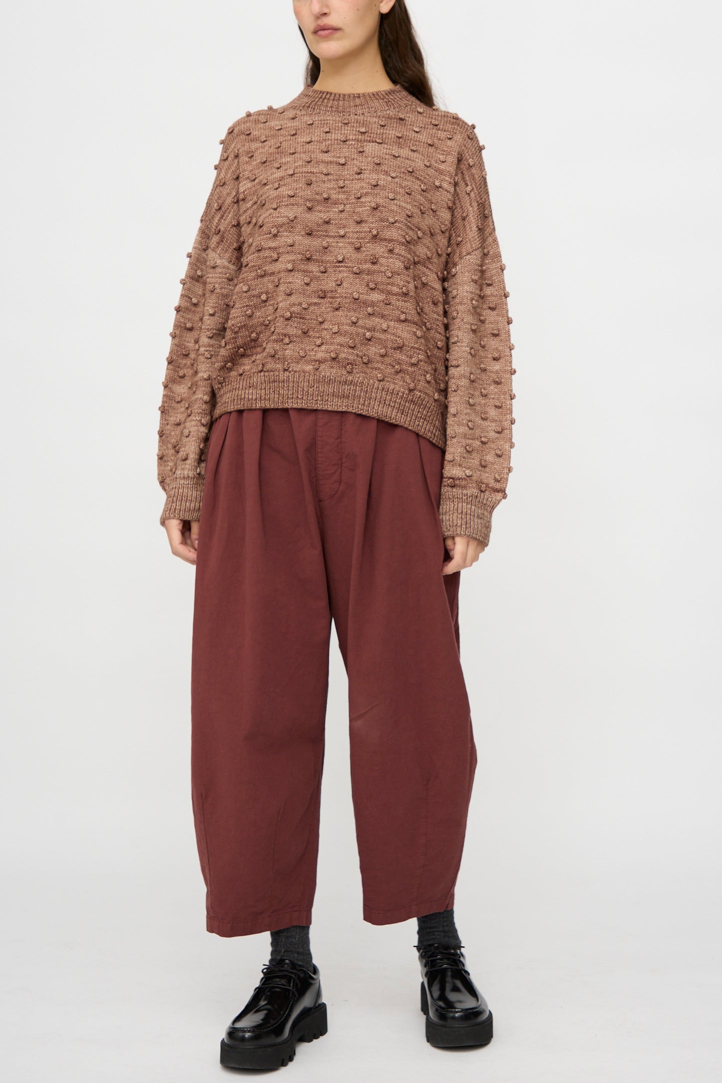 A person wearing the Misha & Puff Original Popcorn Sweater in Affogato, loose maroon pants, dark socks, and black platform shoes stands against a plain light background.
