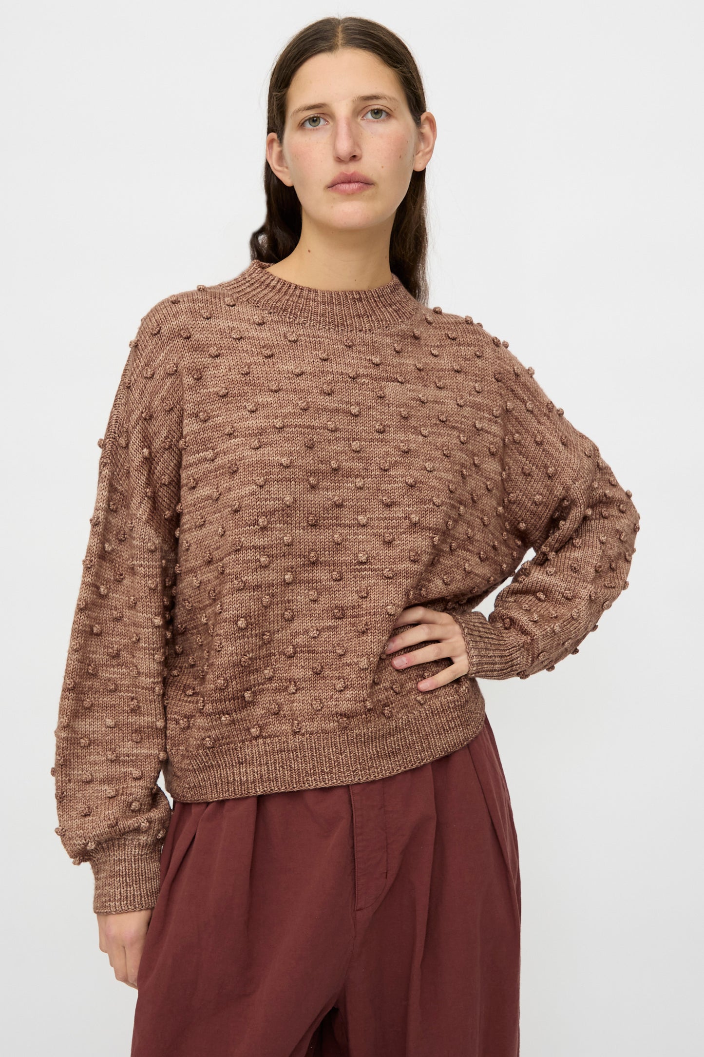 A woman stands against a plain background, wearing the Misha & Puff Original Popcorn Sweater in Affogato and loose maroon pants, with one hand on her hip.