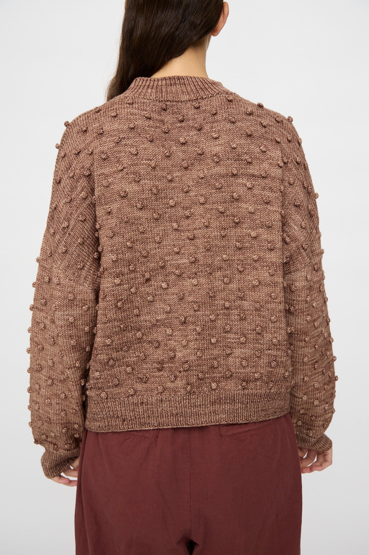 A person faces away, wearing the Misha & Puff Original Popcorn Sweater in Affogato with raised bobble details and maroon pants. Their long, straight hair extends down their back.
