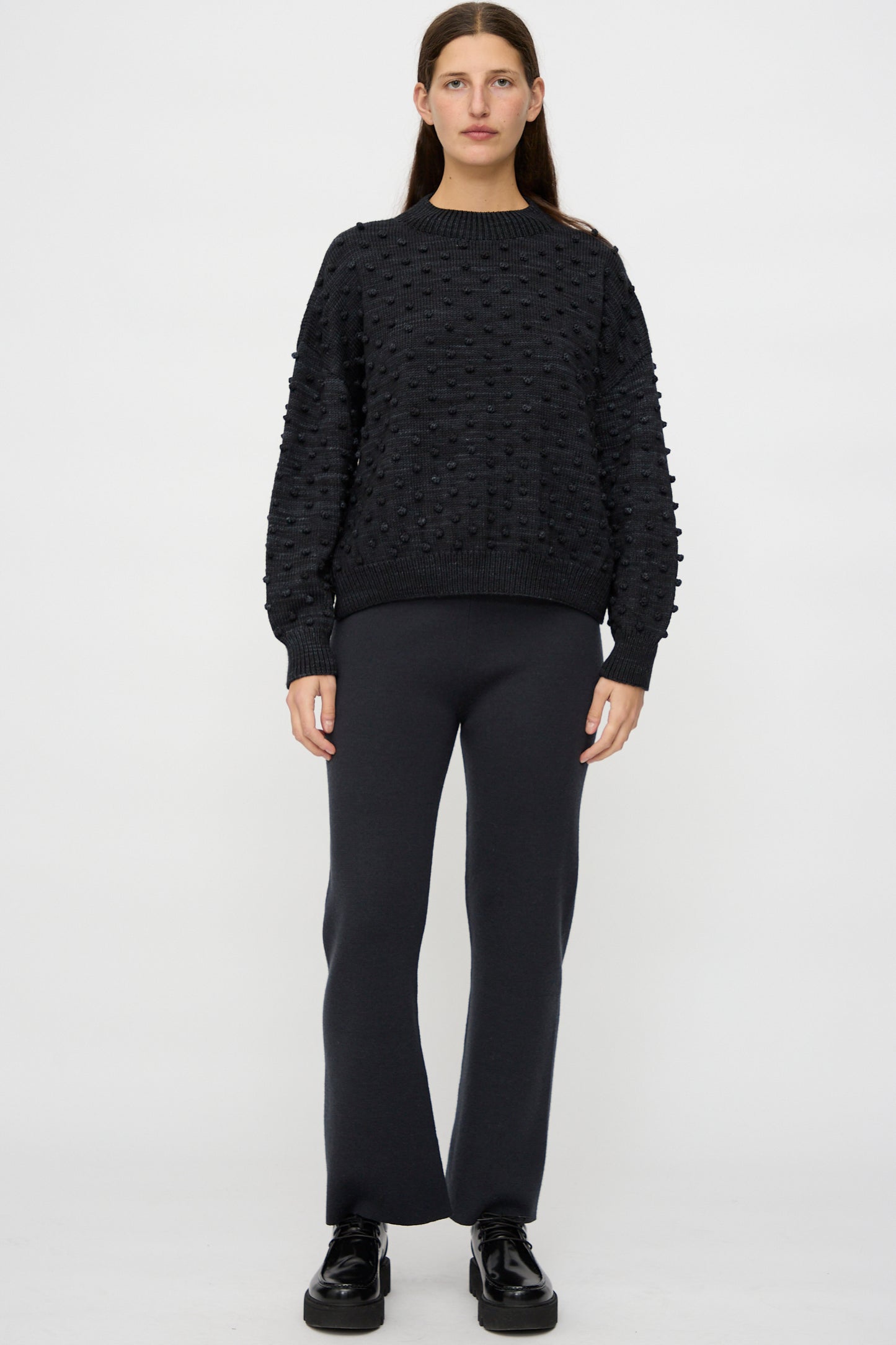 A person stands facing forward against a plain white background, wearing the Misha & Puff Original Popcorn Sweater in Carbon, paired with straight-leg black pants and black shoes.