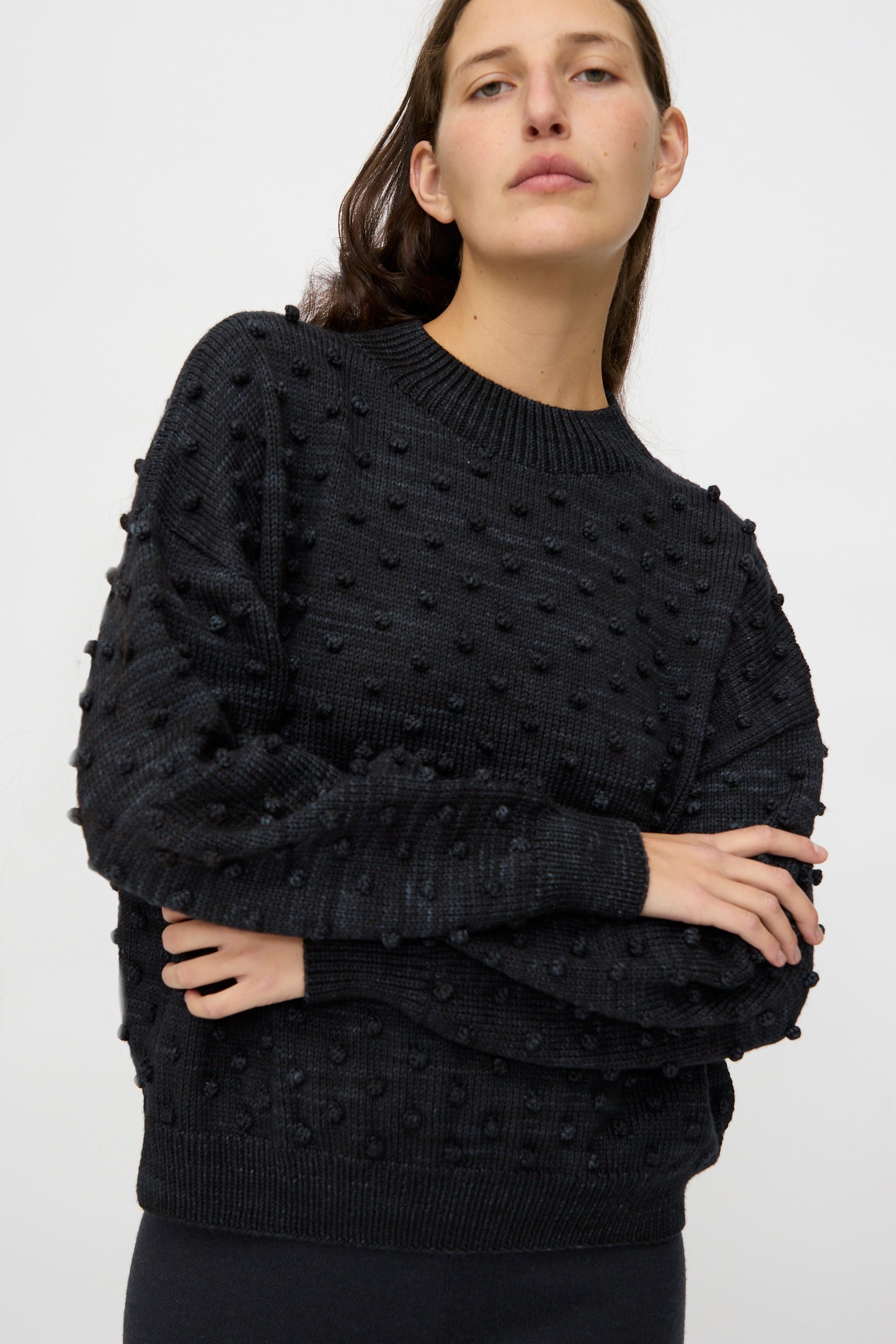 A person with long brown hair wears the Misha & Puff Original Popcorn Sweater in Carbon, featuring raised dots, standing against a plain white background with arms crossed.