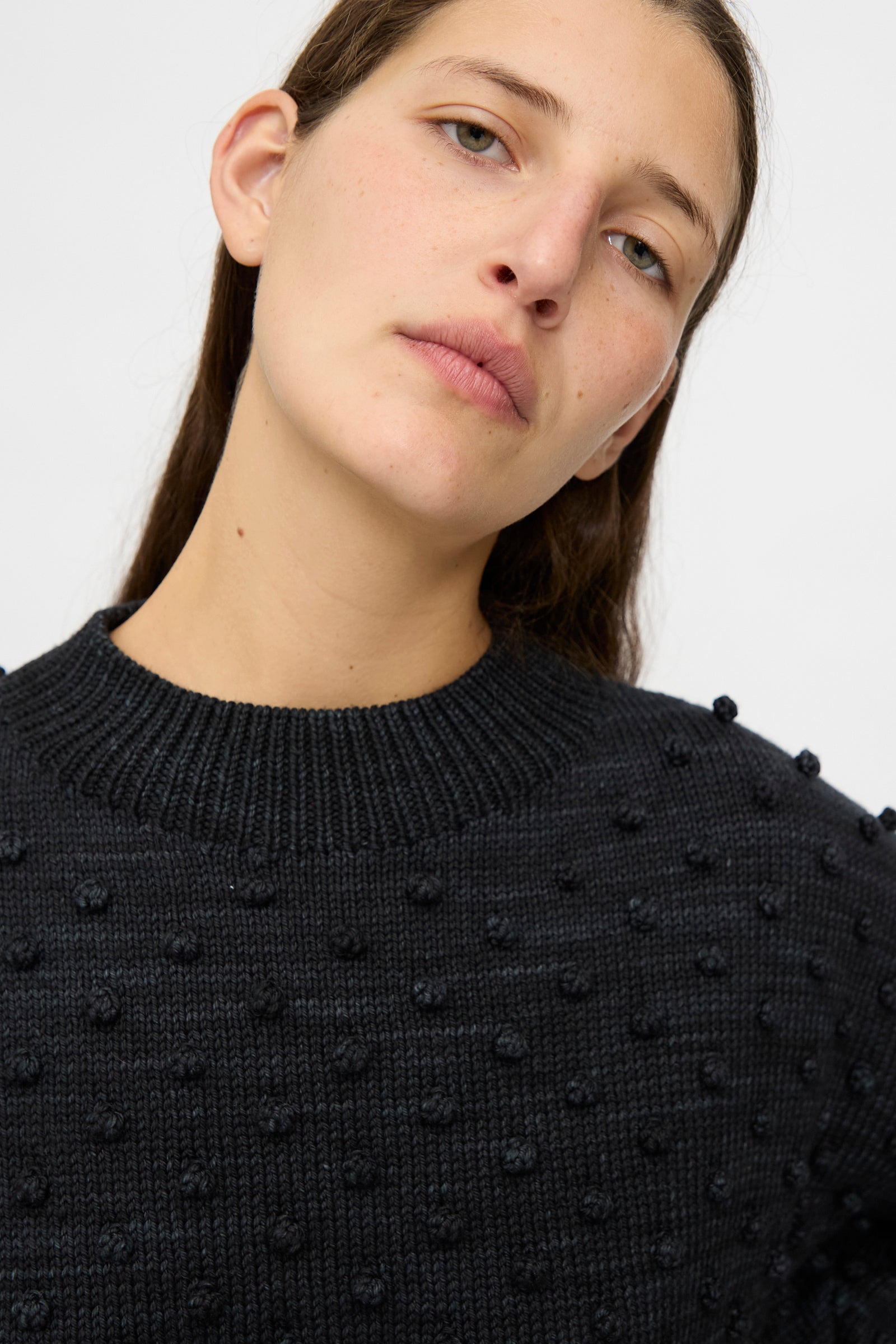 A woman with long brown hair wears the Misha & Puff Original Popcorn Sweater in Carbon, showcasing textured popcorn-stitch dots against a plain light background.