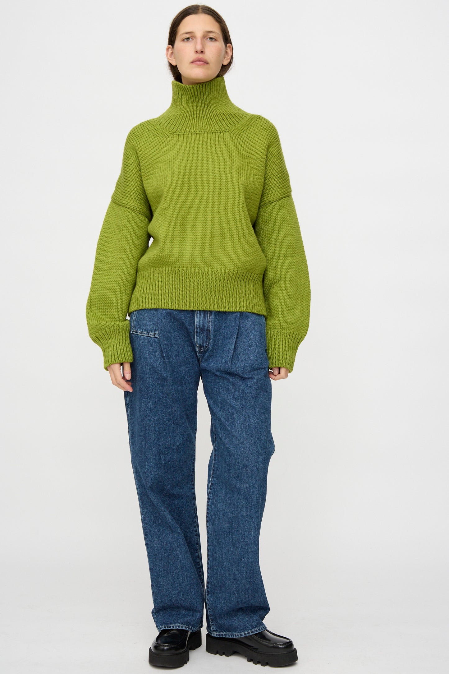 A person stands against a plain background wearing an oversized Misha & Puff Turtleneck Sweater in Matcha, loose blue jeans, and black shoes.