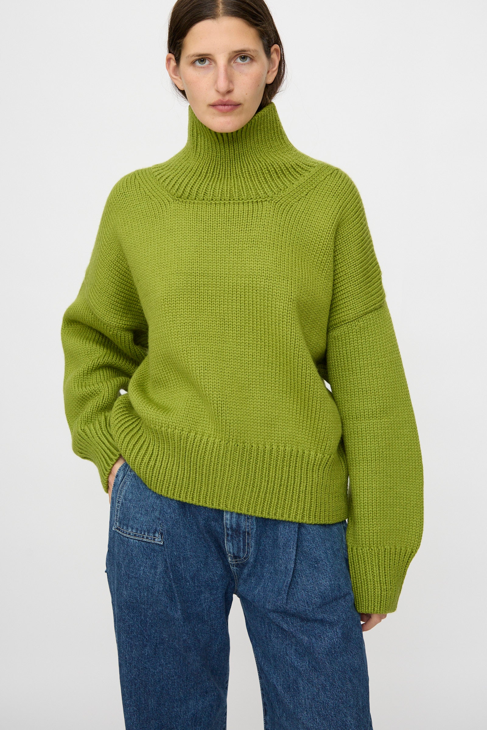 A person wears the Misha & Puff Turtleneck Sweater in Matcha, styled in an oversized fit with blue jeans, standing against a plain white background.