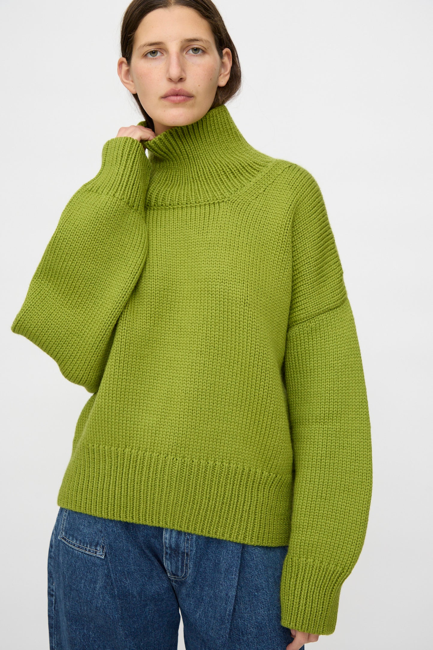 A person wears the Misha & Puff Turtleneck Sweater in Matcha, featuring a chunky, oversized fit, paired with blue jeans and standing against a plain white background.