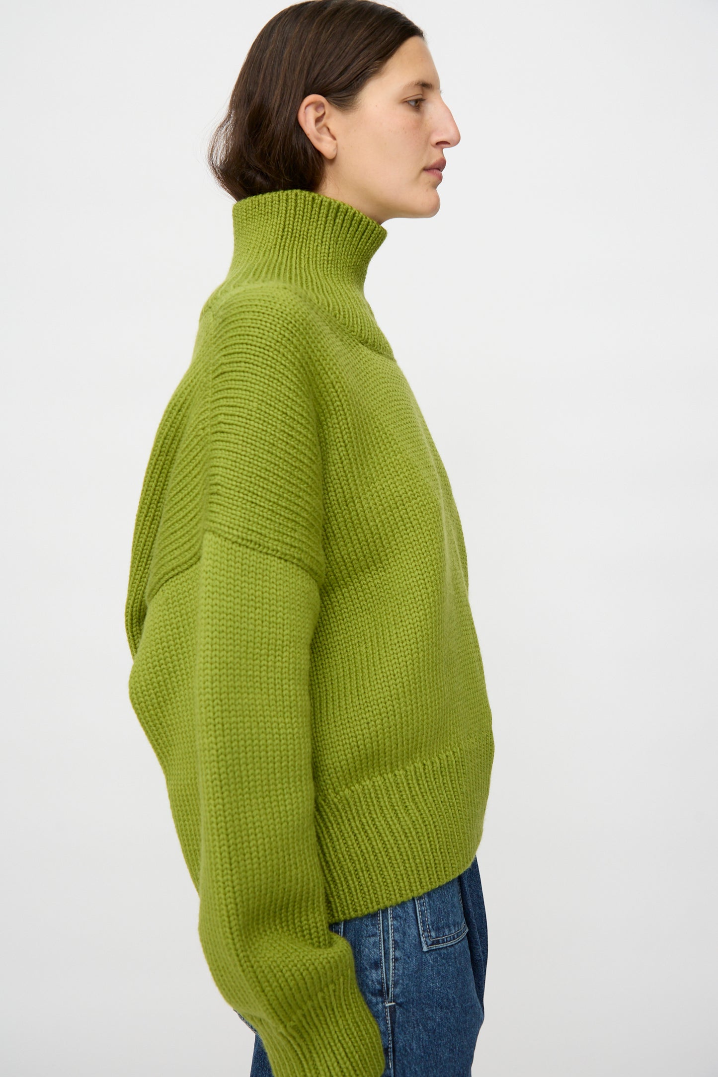 A person in an oversized fit, Misha & Puff Turtleneck Sweater in Matcha and blue jeans stands sideways against a plain light background.