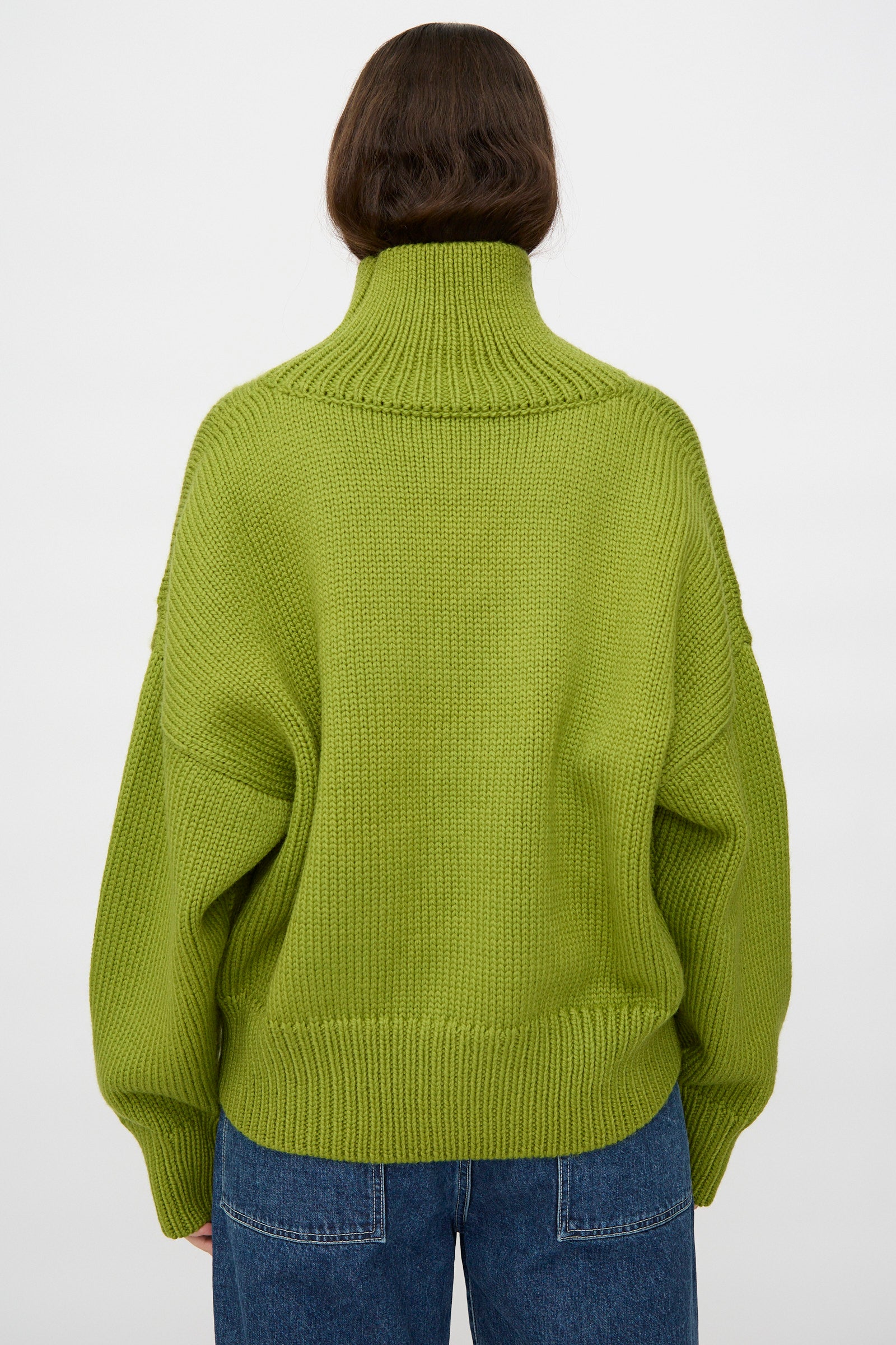 A person with long brown hair wears the Misha & Puff Turtleneck Sweater in Matcha and blue jeans, facing away from the camera against a plain white background.