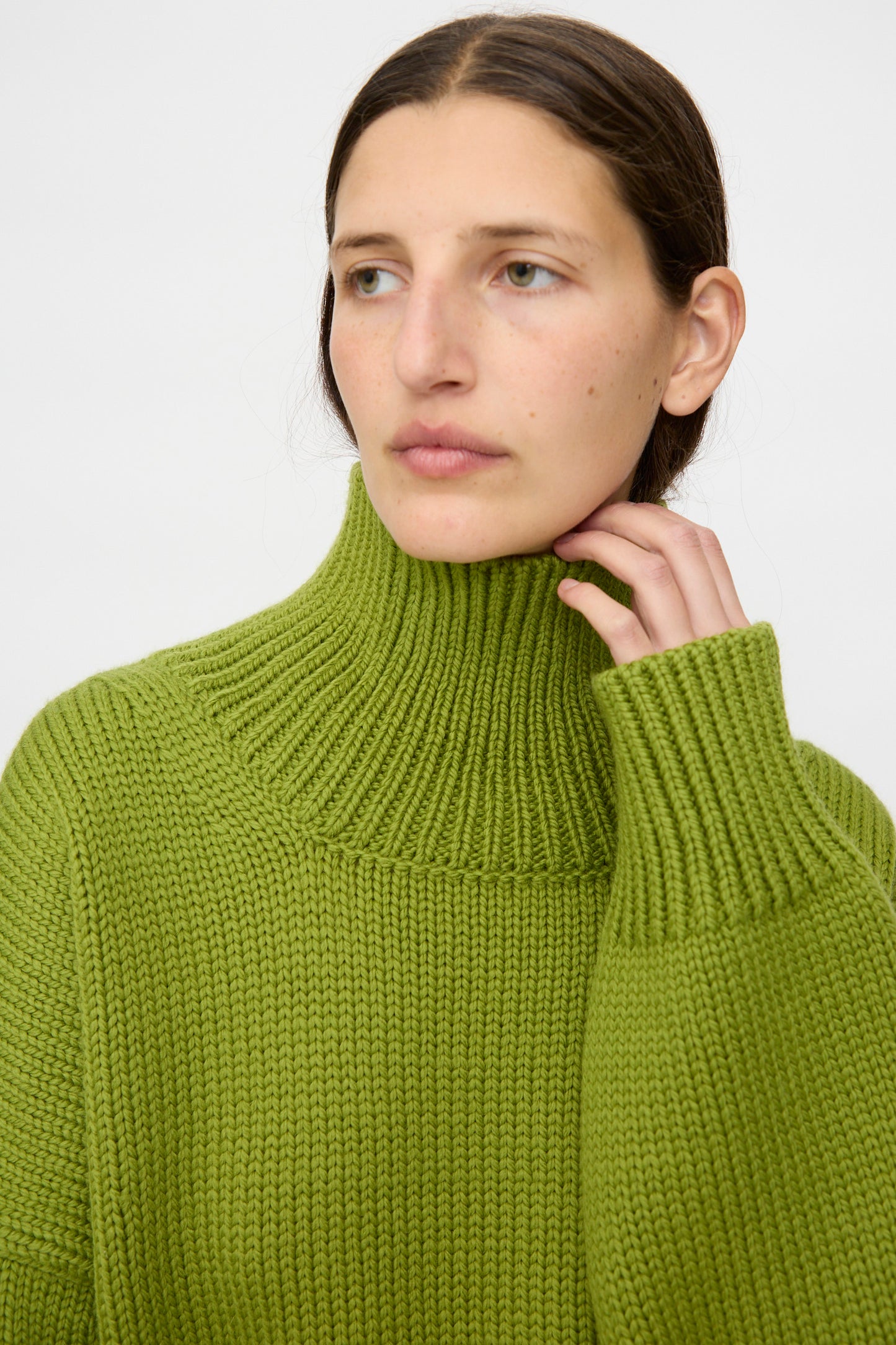 A person with long brown hair, wearing the Misha & Puff Turtleneck Sweater in Matcha with an oversized fit, looks slightly to the side and touches the collar.