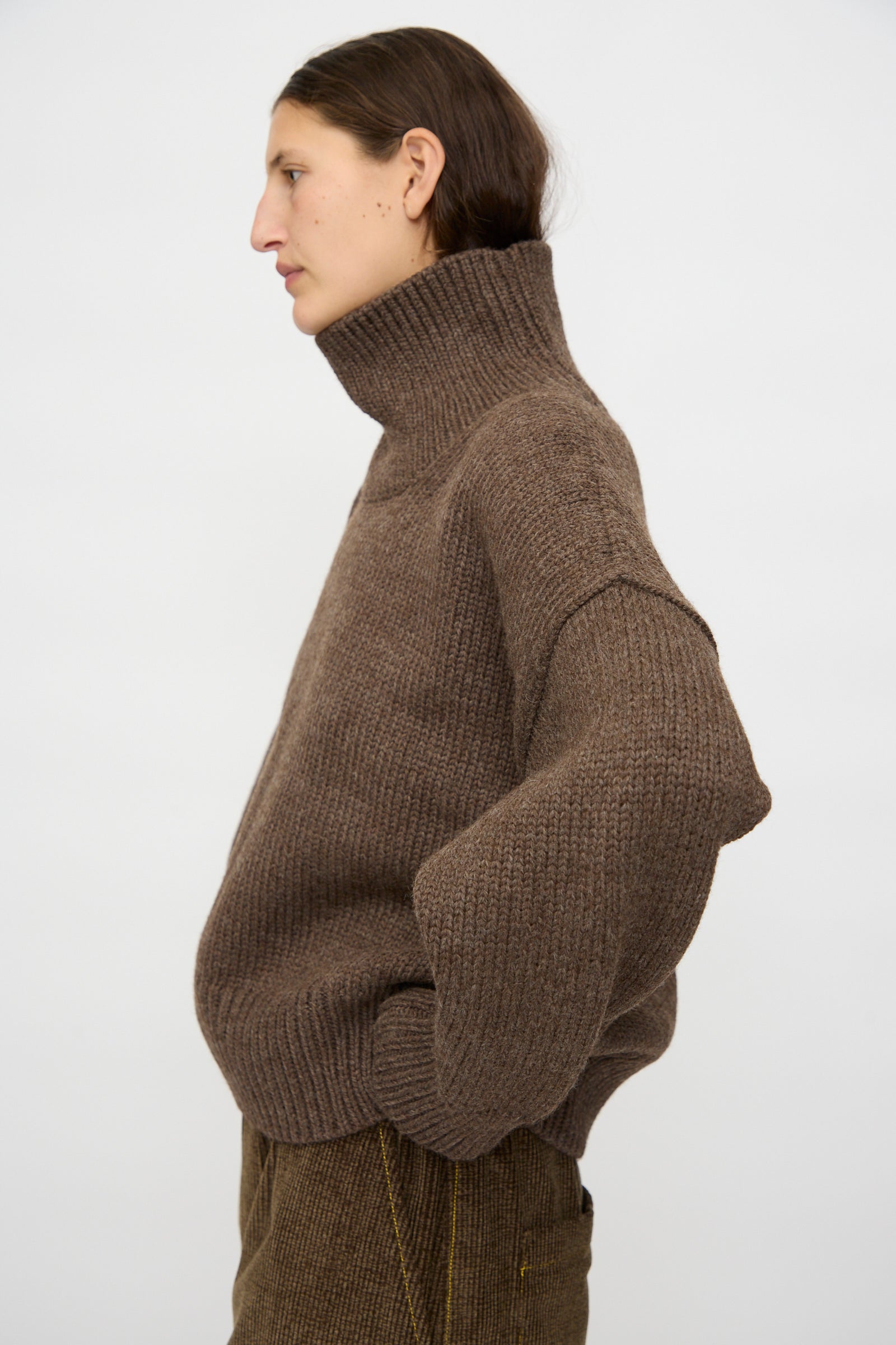 A person with long brown hair, wearing the Misha & Puff Turtleneck Sweater in Oak and brown pants, stands in profile against a plain white background.